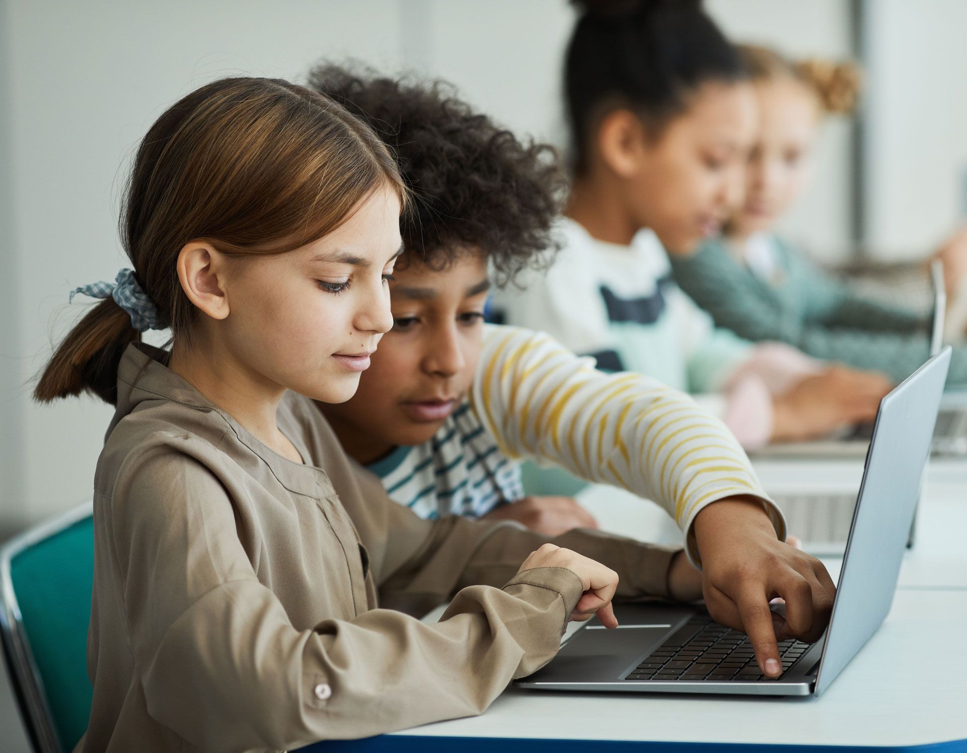 Children working together on a laptop at a classroom table, with one pointing at the screen.