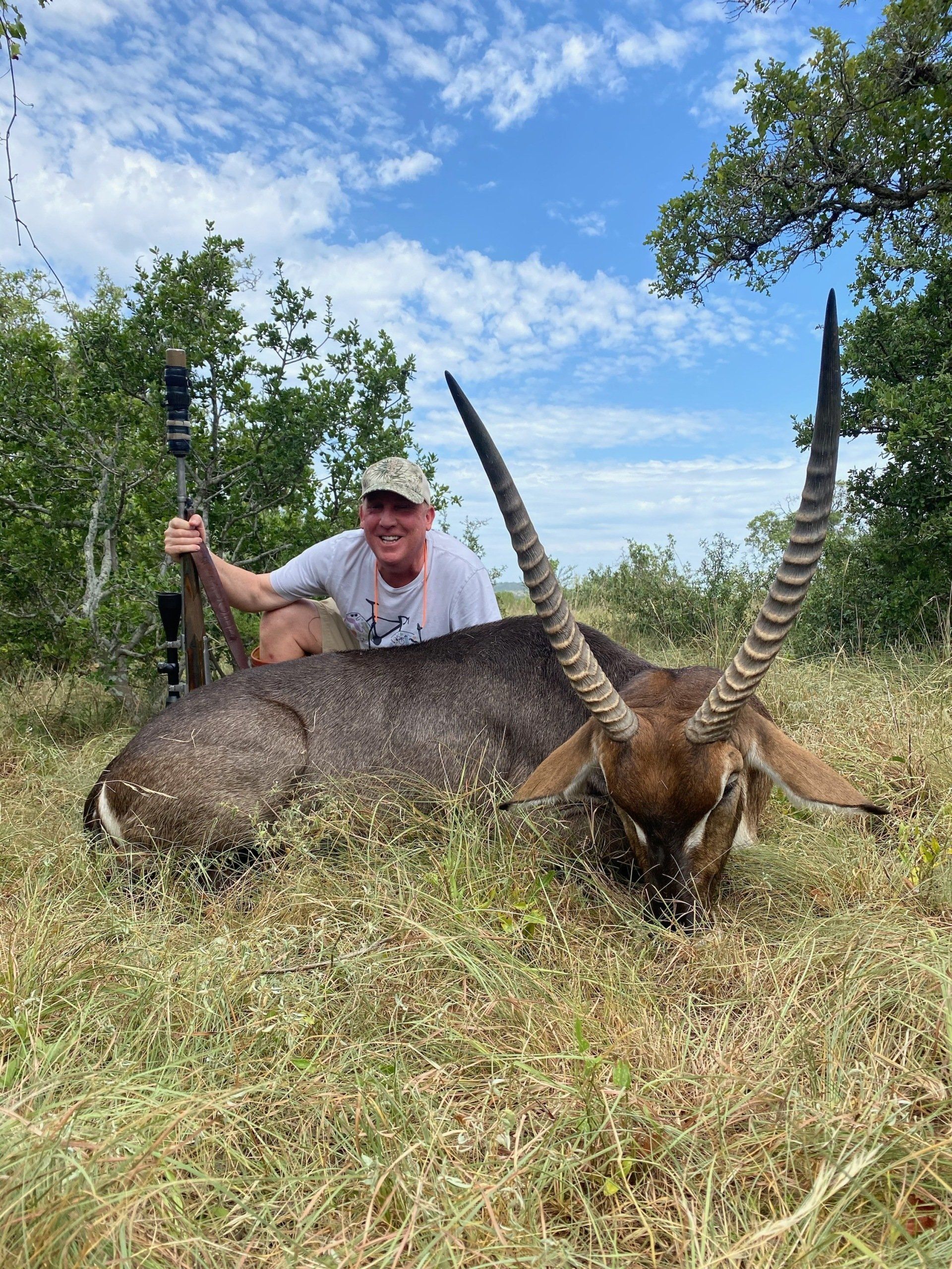 Man kneeling next to a dead waterbuck in grassy field, holding rifle. Blue sky.