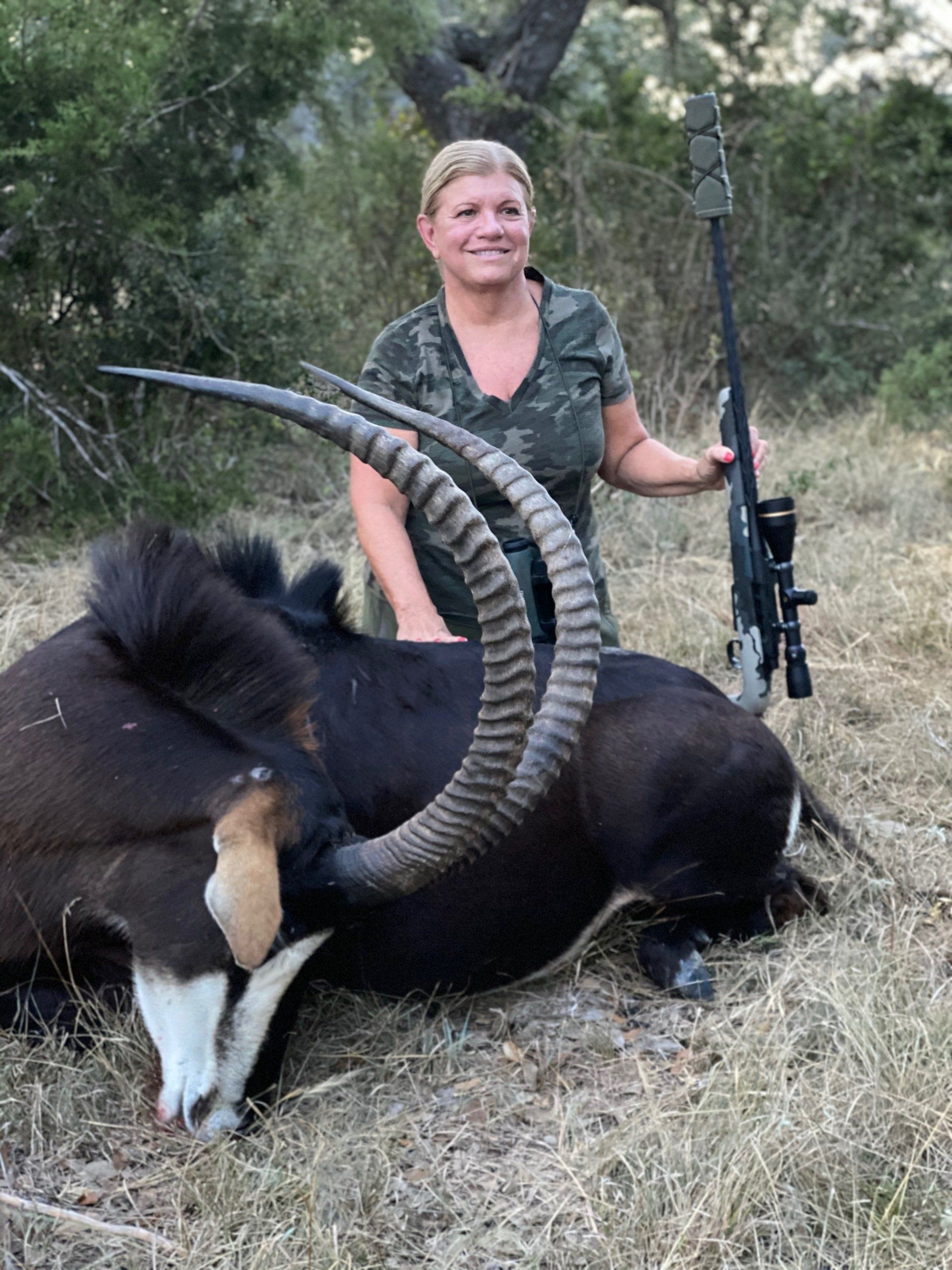 Woman smiles, posing with a dead sable antelope, holding a rifle in a grassy area.