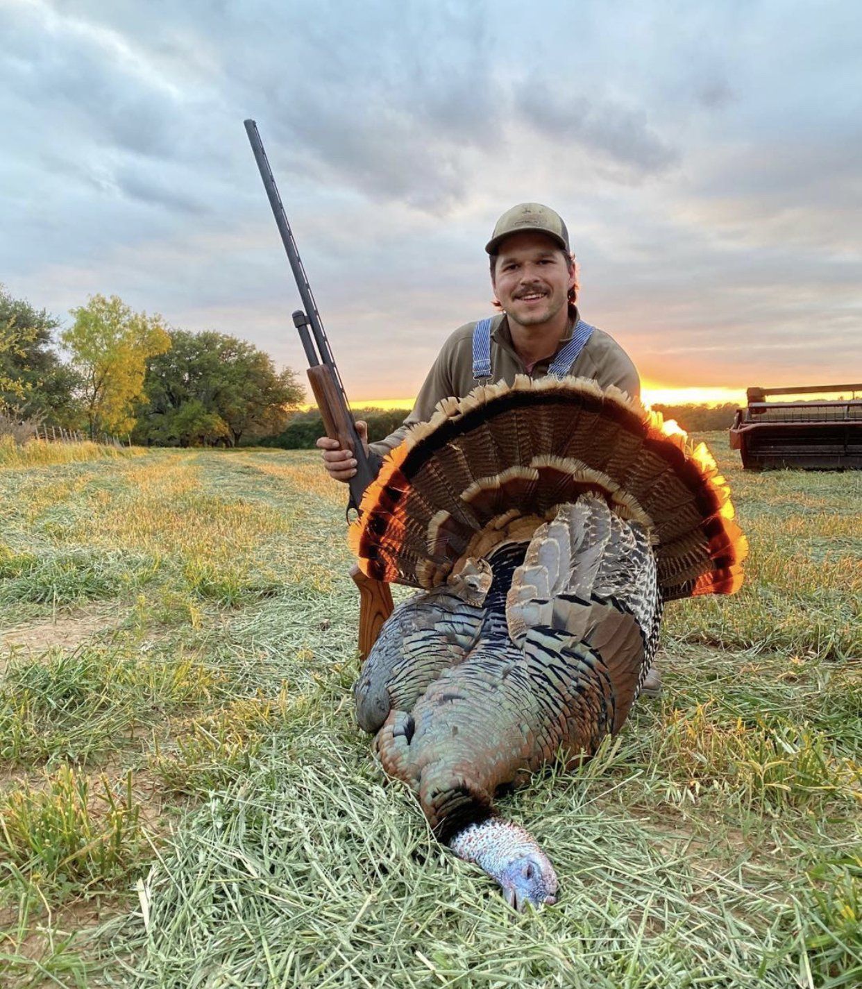 Man holding a turkey he has hunted, with a shotgun. Outdoors, sunset setting, grassy field.