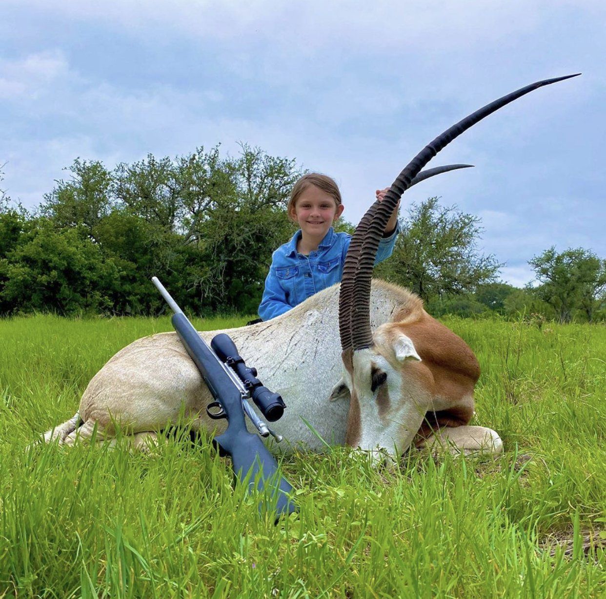 Young girl posing with a recently hunted scimitar-horned oryx in a grassy field; rifle visible.