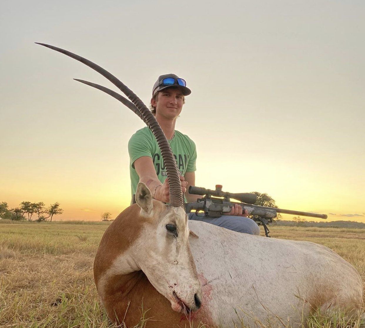 Man with a rifle posing with a dead Oryx in a field at sunset.