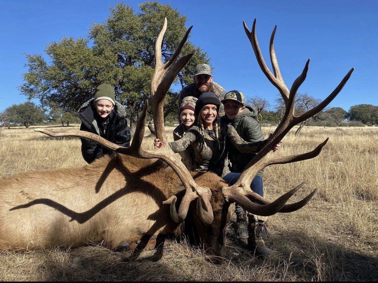 Family poses with a large elk they hunted outdoors. People are smiling, some in camouflage, in a sunny field.