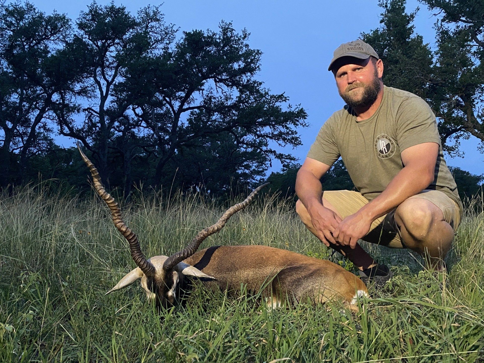 Man kneels beside an antelope he hunted. He wears a hat and khaki shorts in a grassy field.