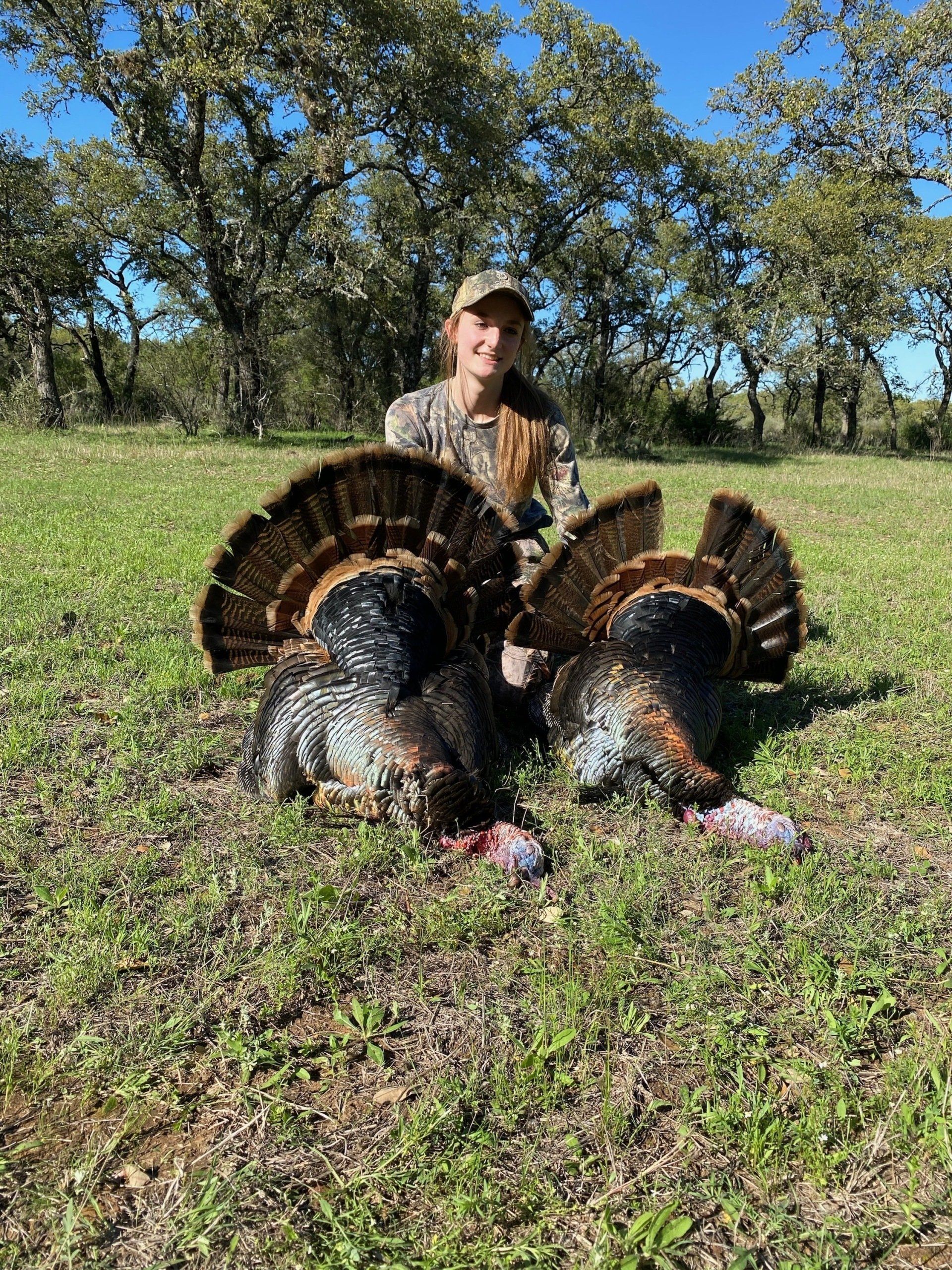 Woman in camouflage with two dead turkeys on a grassy field, smiling.
