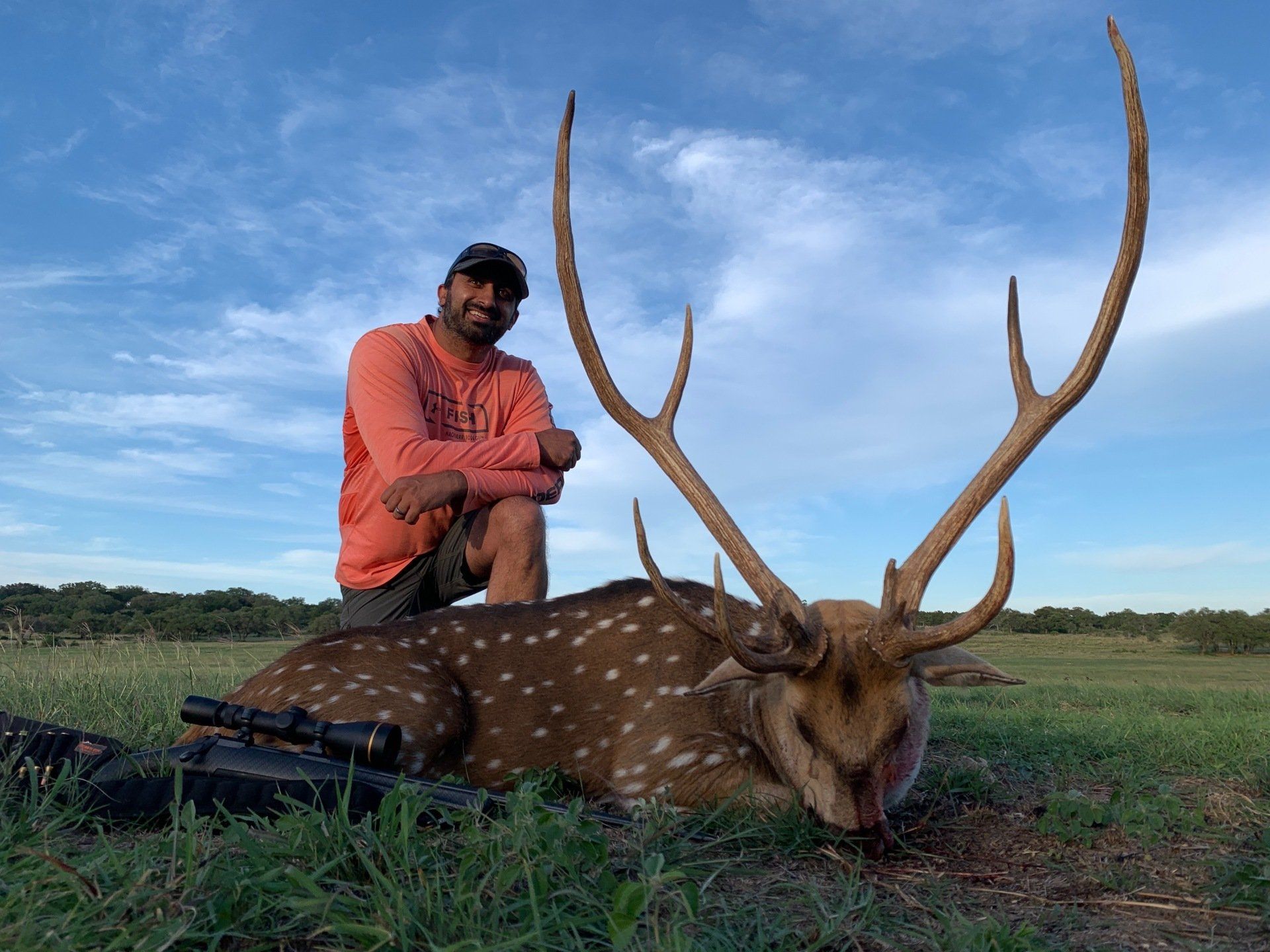 Man kneels beside a spotted deer with large antlers in a grassy field under a cloudy sky.