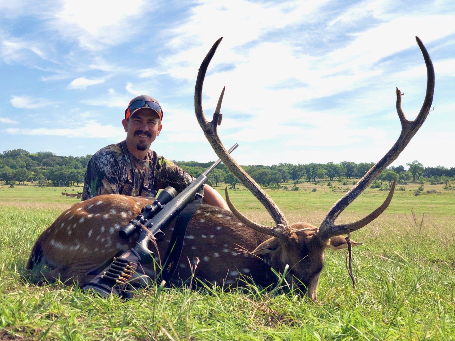 Man in camouflage smiles next to a spotted deer with large antlers, rifle on deer, grassy field.