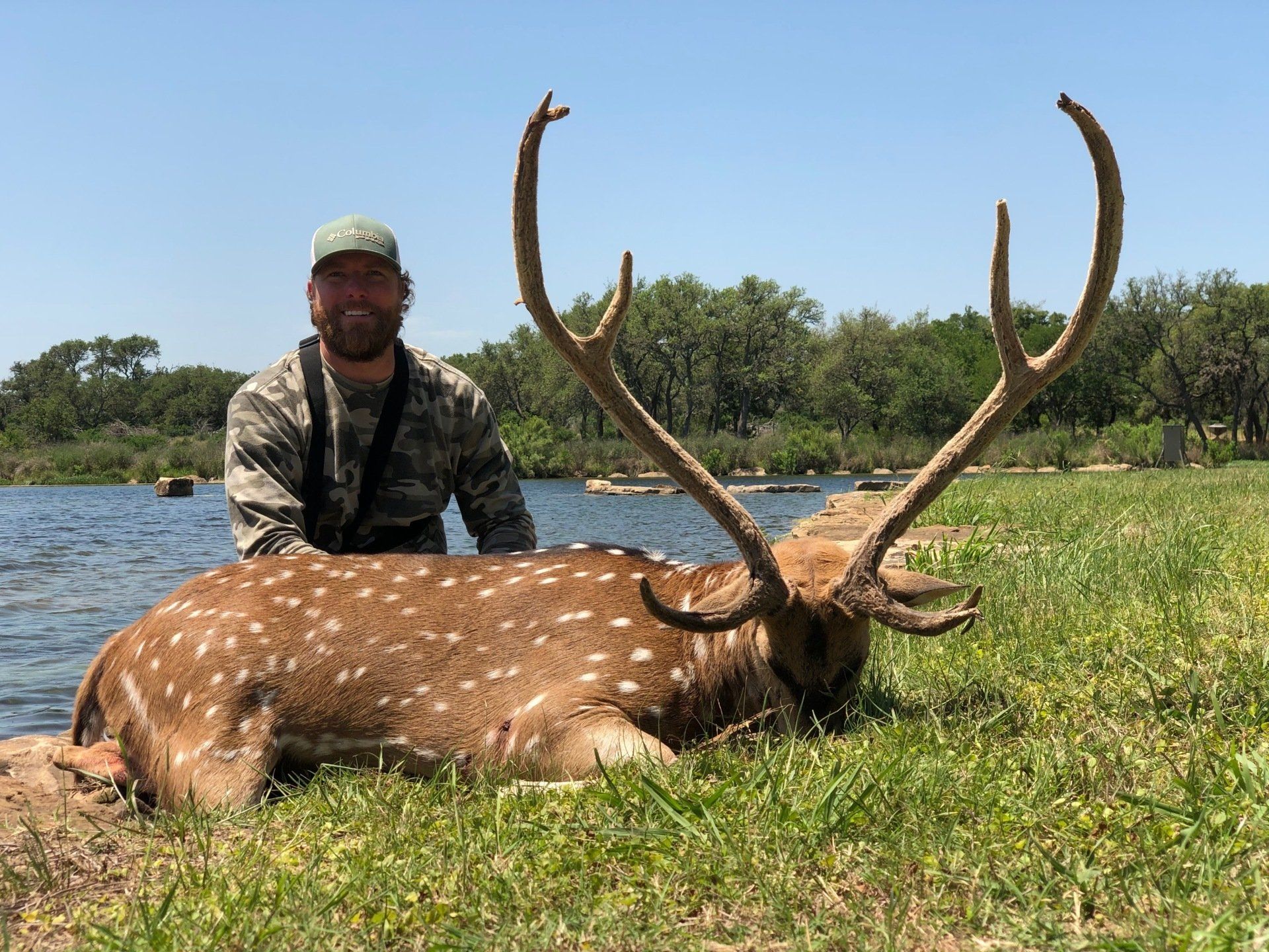 Man in camo next to a spotted Axis deer with large antlers, by a body of water, smiling.