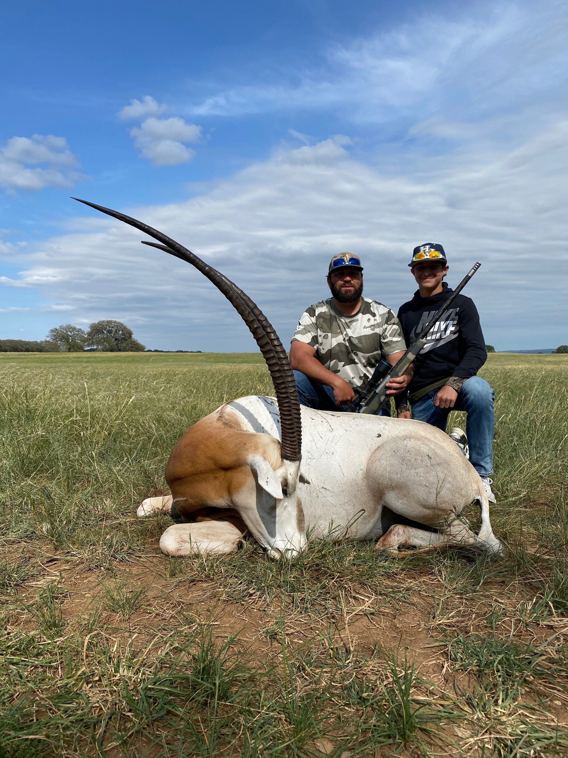 Two men pose with an oryx in a field; one holds a rifle. Blue sky, green grass, and brown earth.