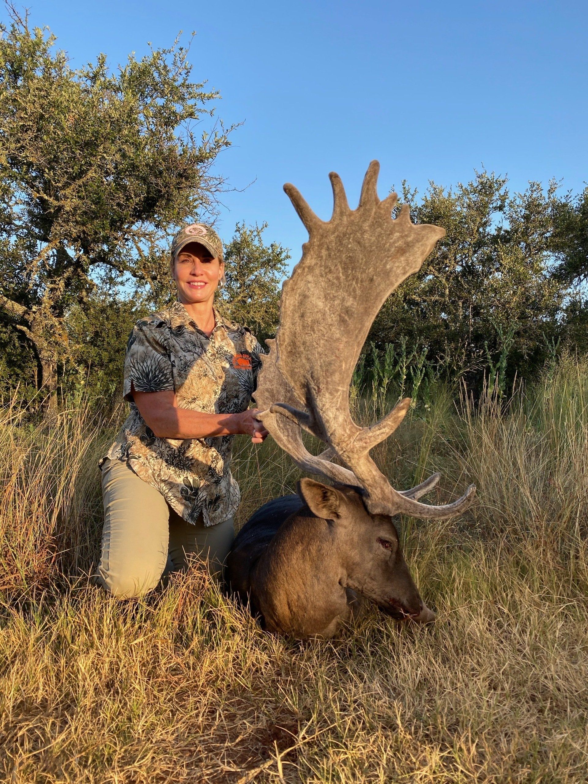 Woman kneels next to a large deer, showing the animal's impressive antlers. They are outdoors, in grassy area.