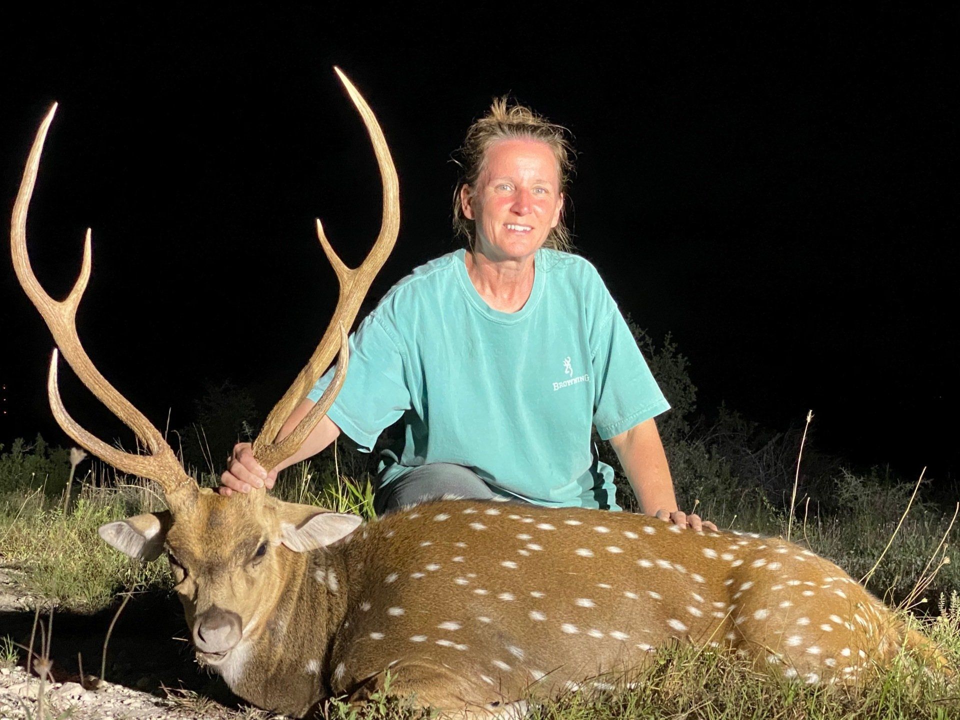 Woman kneels, smiling, beside a spotted deer with large antlers, outdoors at night.