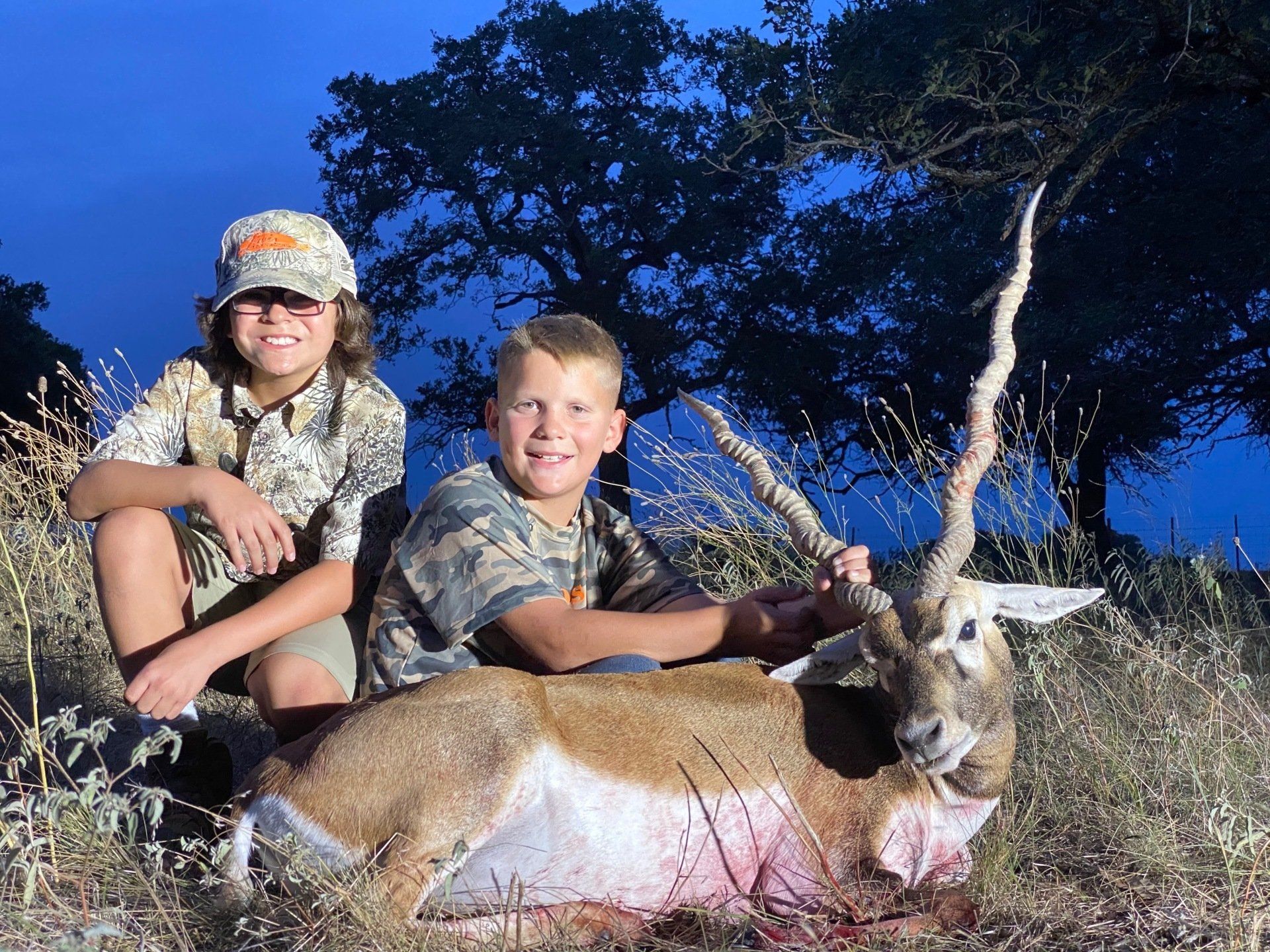 Two boys in camo with a blackbuck. One boy smiles. Outdoors at dusk.