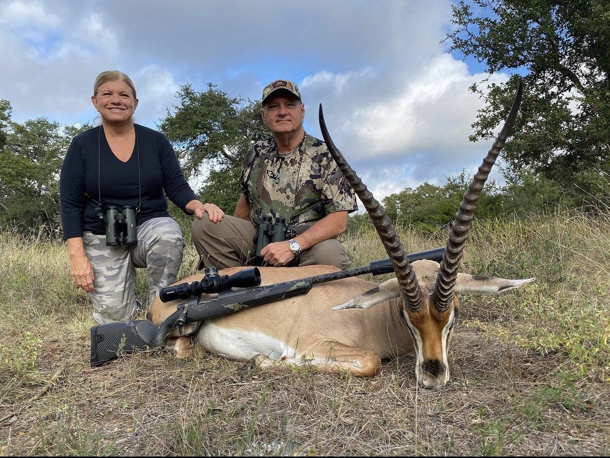 Two people kneeling next to a dead antelope, outdoors. The woman smiles, the man is looking at the camera.