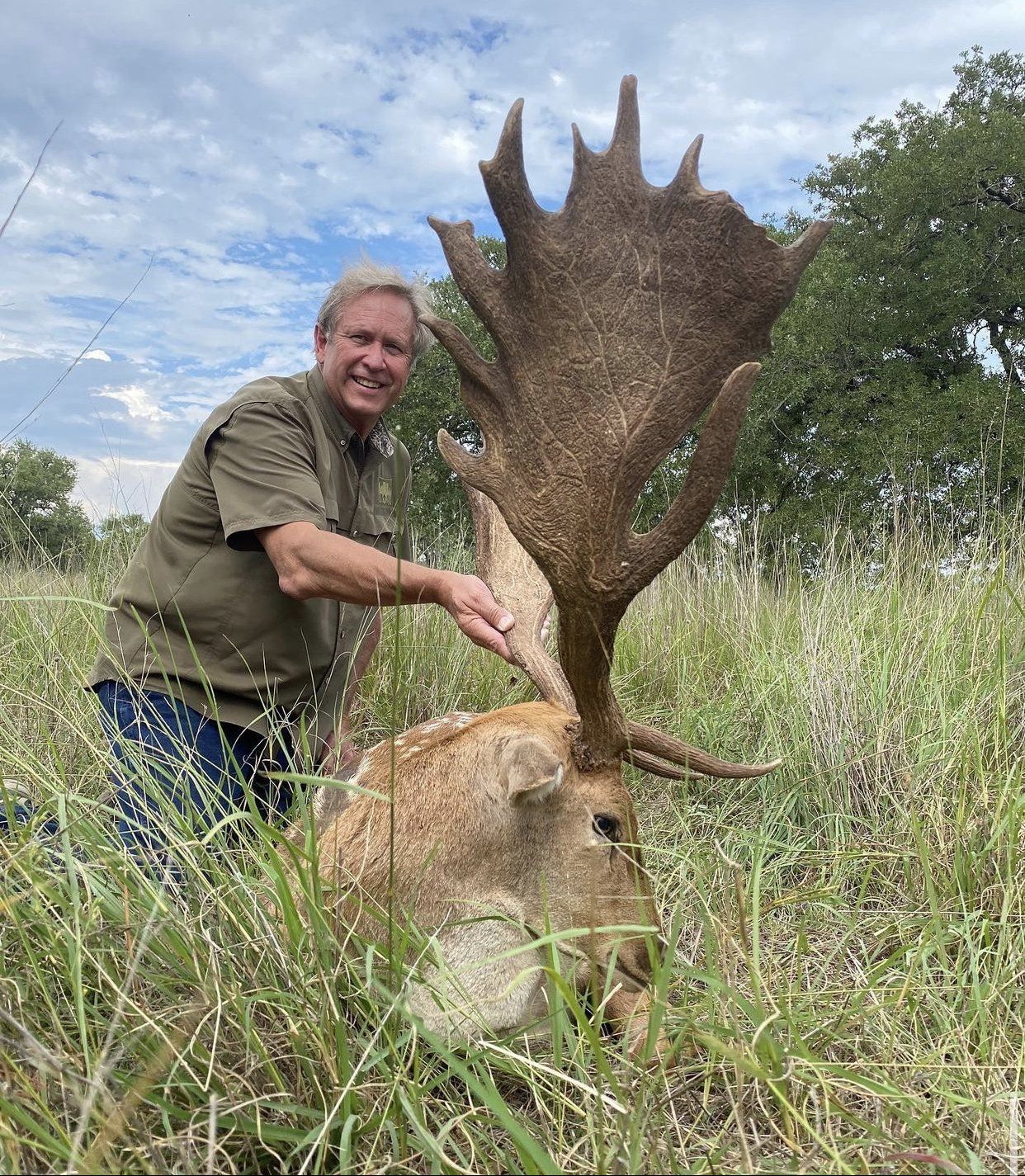 Man kneeling beside a large deer with impressive antlers in tall grass. The man smiles.