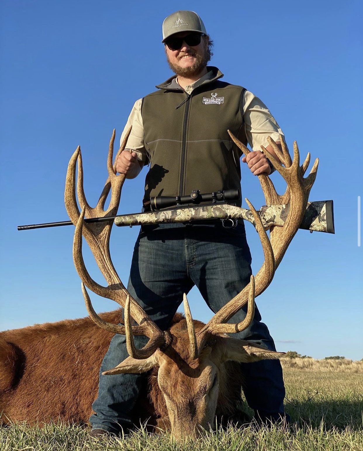 Man smiles, holding rifle across large deer antlers; green vest, blue jeans, sunny field.