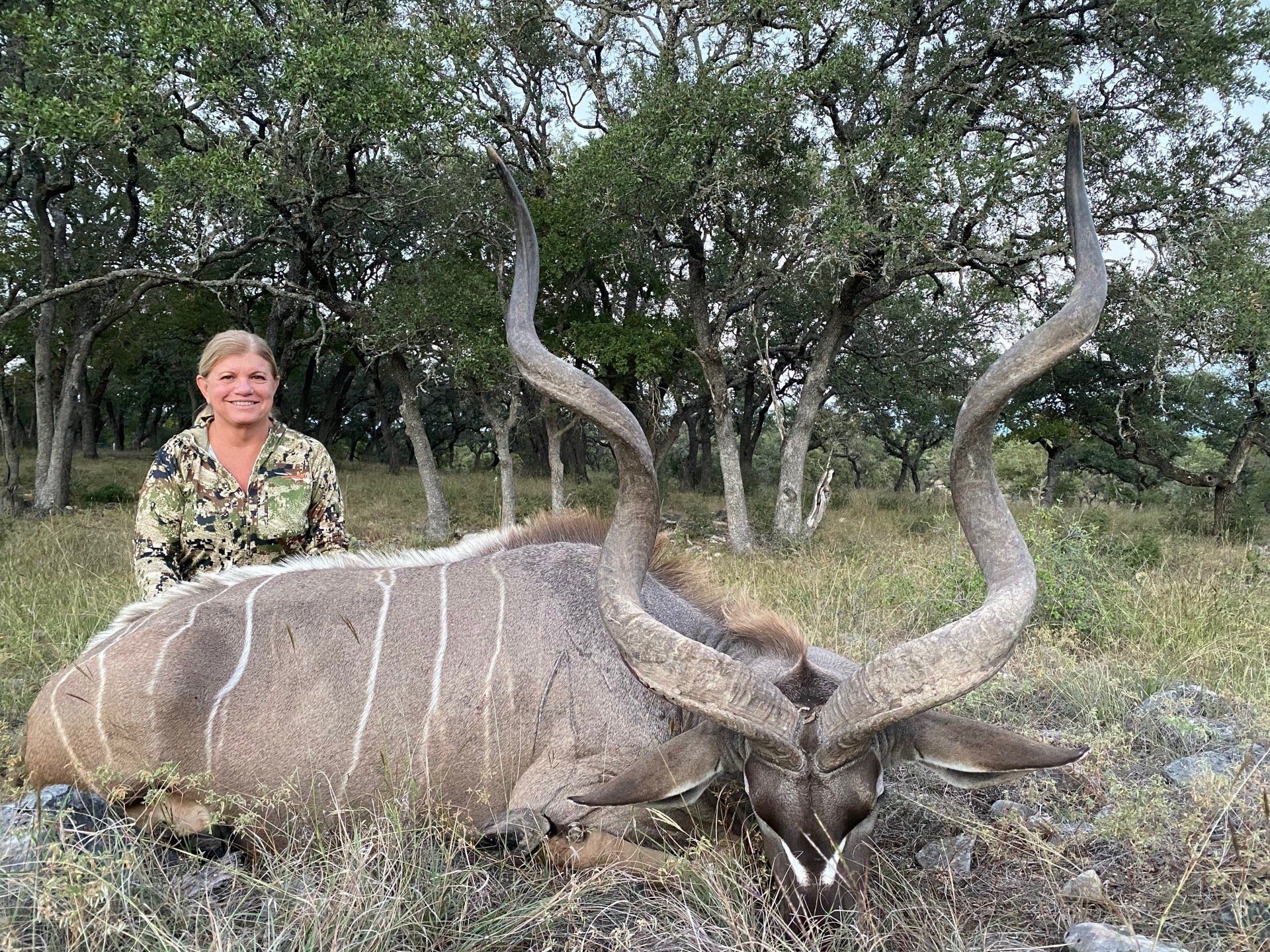 Woman in camo stands next to a dead kudu with large, curved horns in a grassy area.