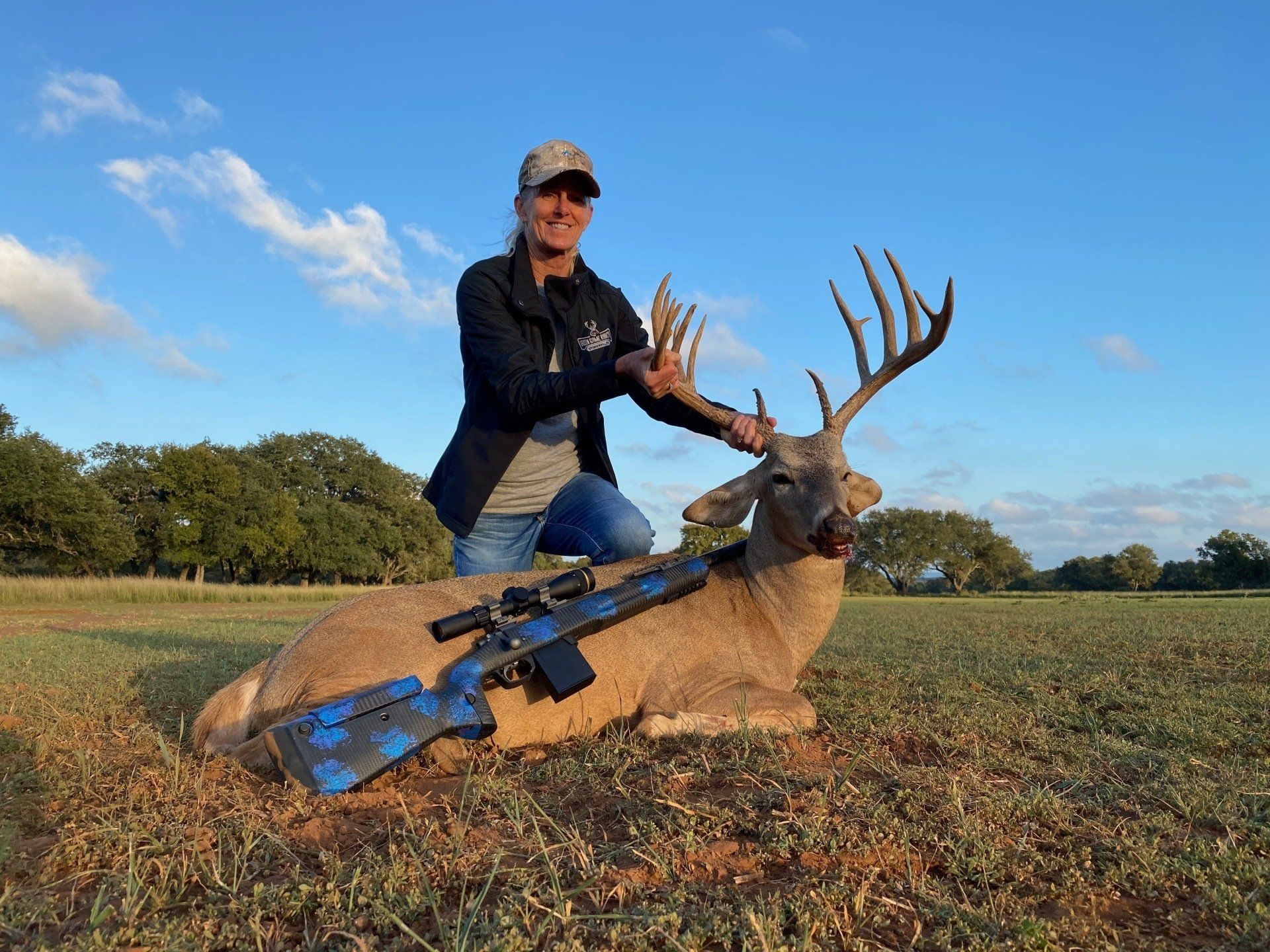 Woman kneels beside a harvested deer in a field, holding its antlers. A blue and black rifle is beside the deer.