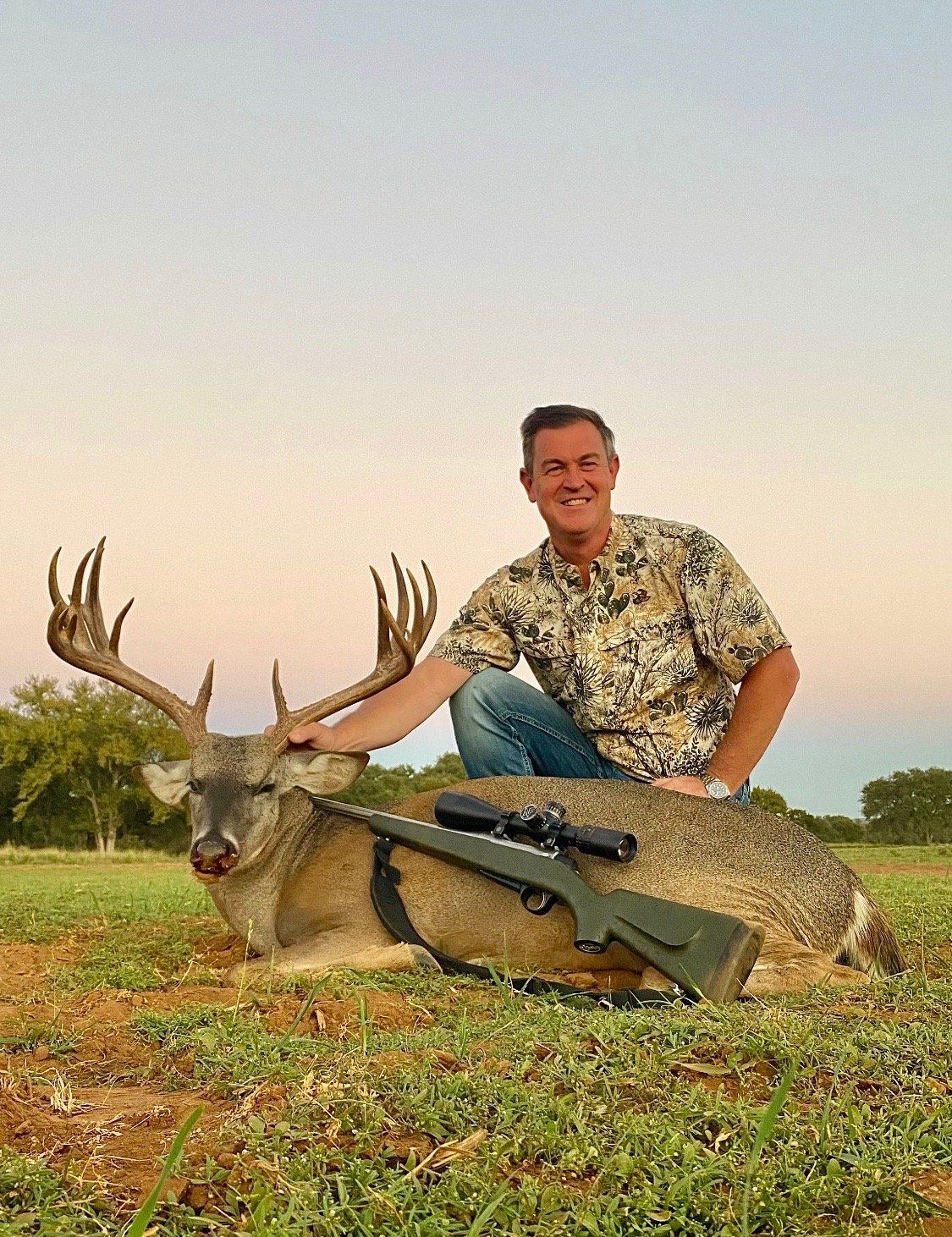 Man kneels beside a dead deer with large antlers, rifle resting on it. Outdoors, setting sun, green grass.