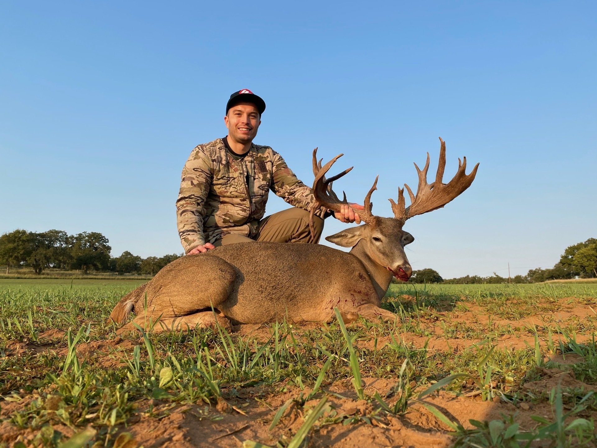 Man in camo kneels next to a large buck in a field, holding the antlers. Blue sky.