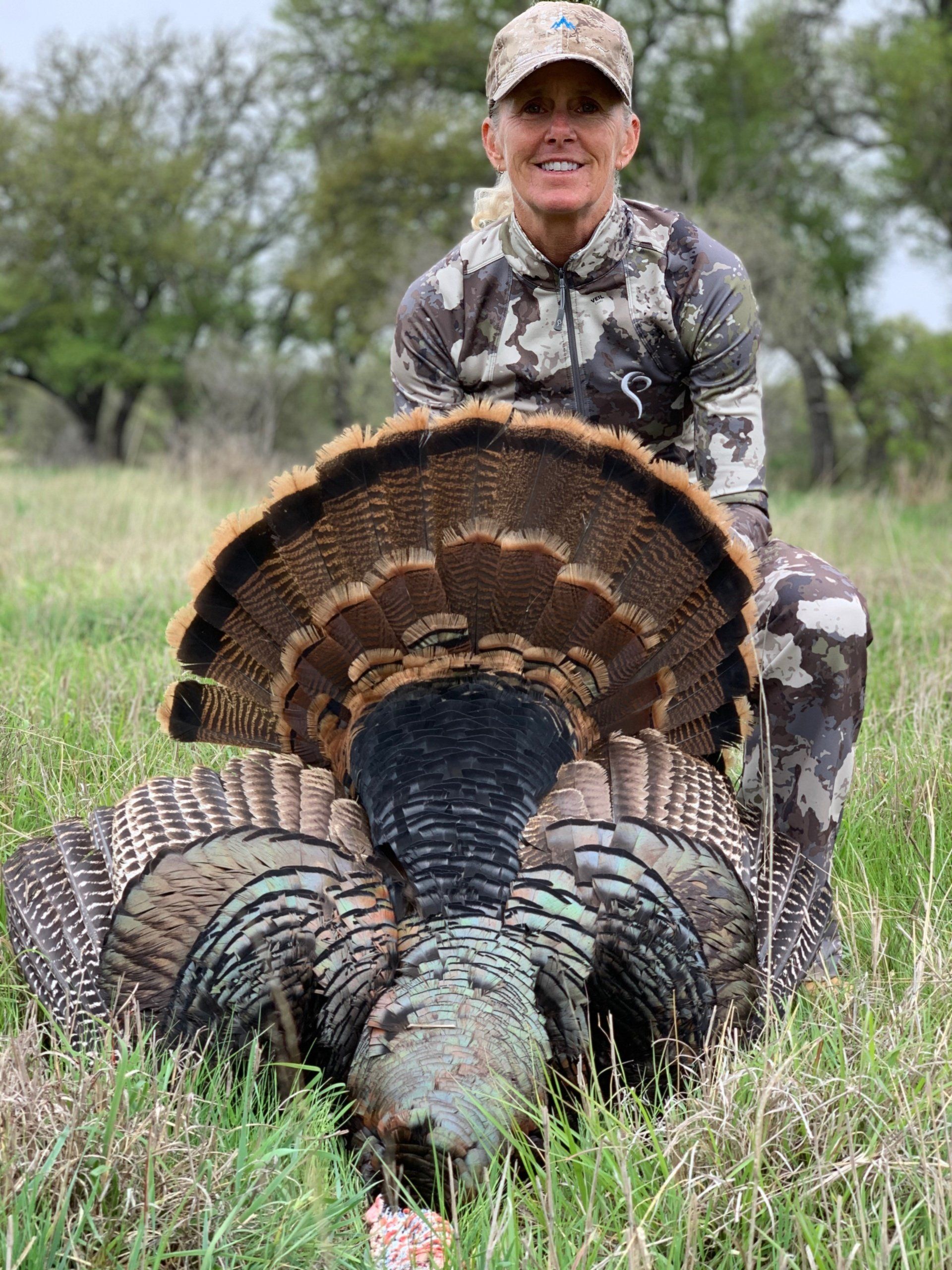 Woman in camouflage kneels proudly with a large harvested wild turkey, field setting.