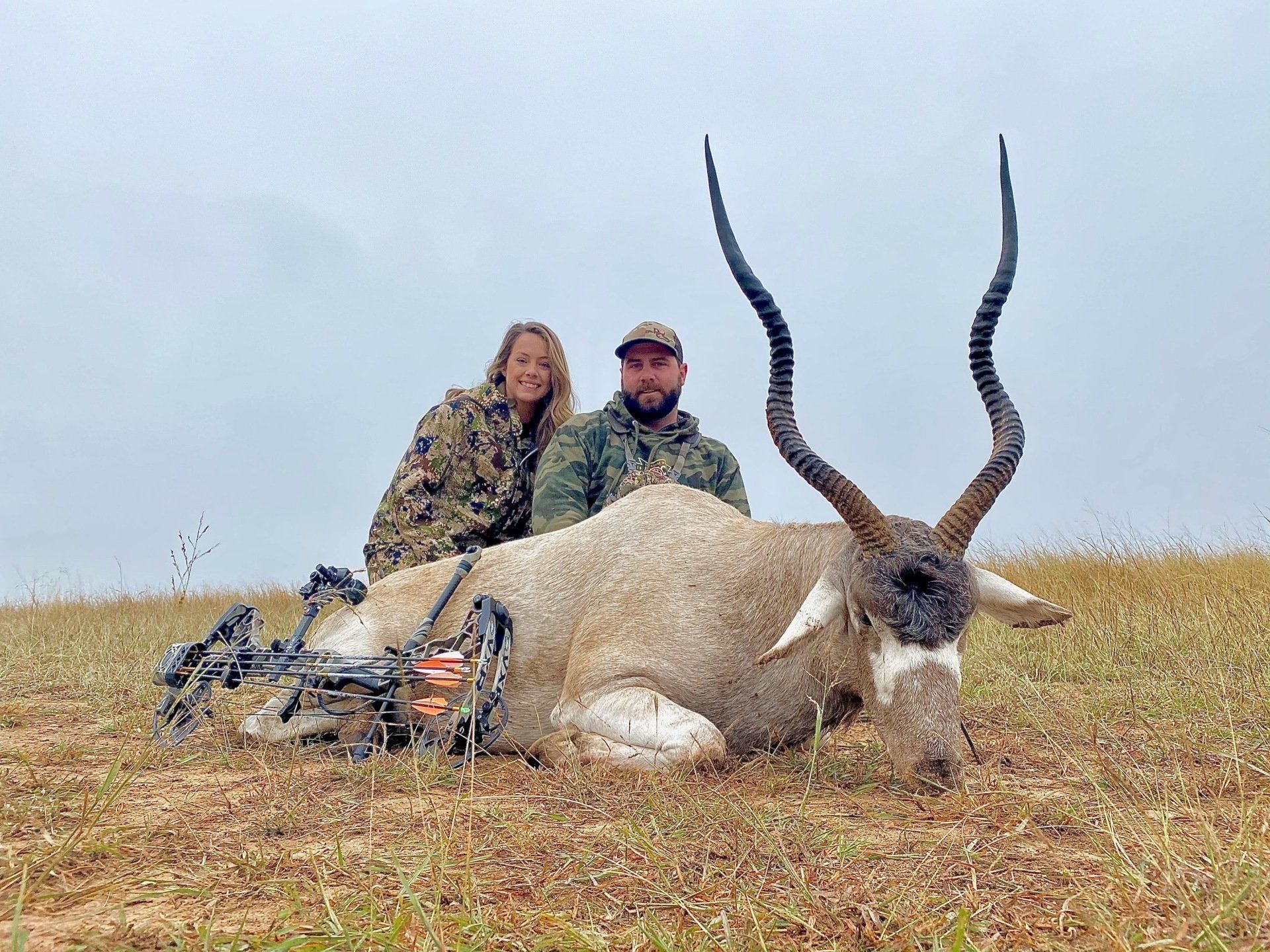 Two people pose with a dead Addax, bow visible. Overcast sky, dry grassy field.