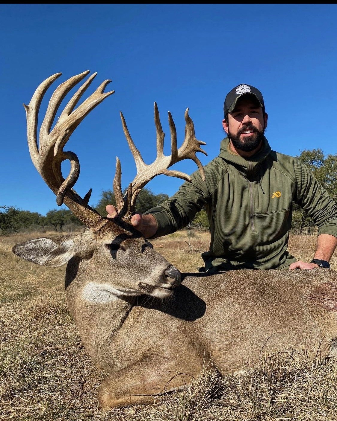 Man with a beard poses with a large buck, holding its antlers, in a field on a sunny day.