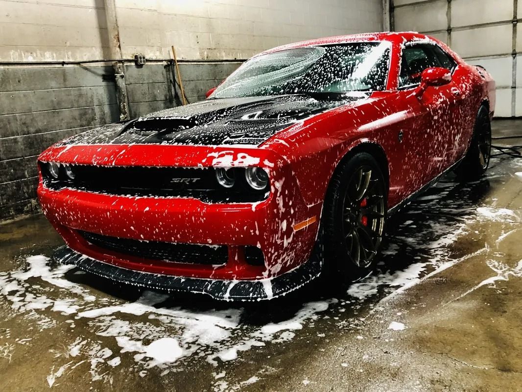 Red sports car being washed in a wet garage, covered in soap foam and water splashes.