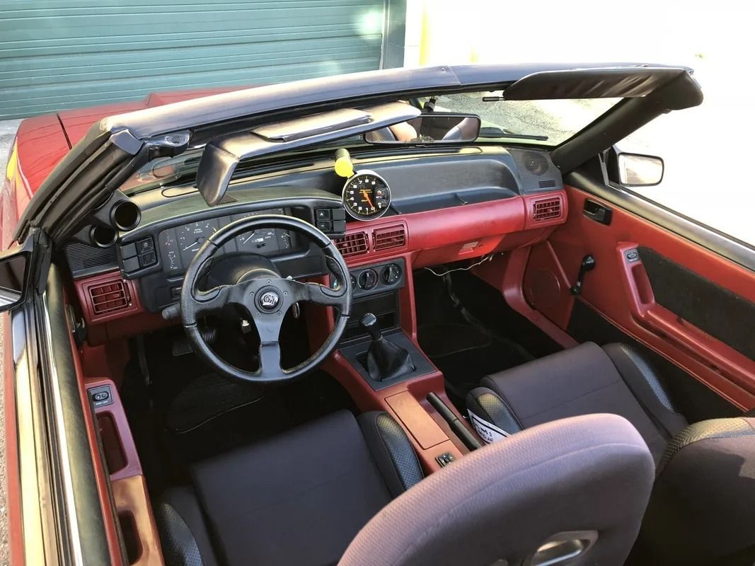Classic car interior with red dashboard, black steering wheel, and open roof by the water