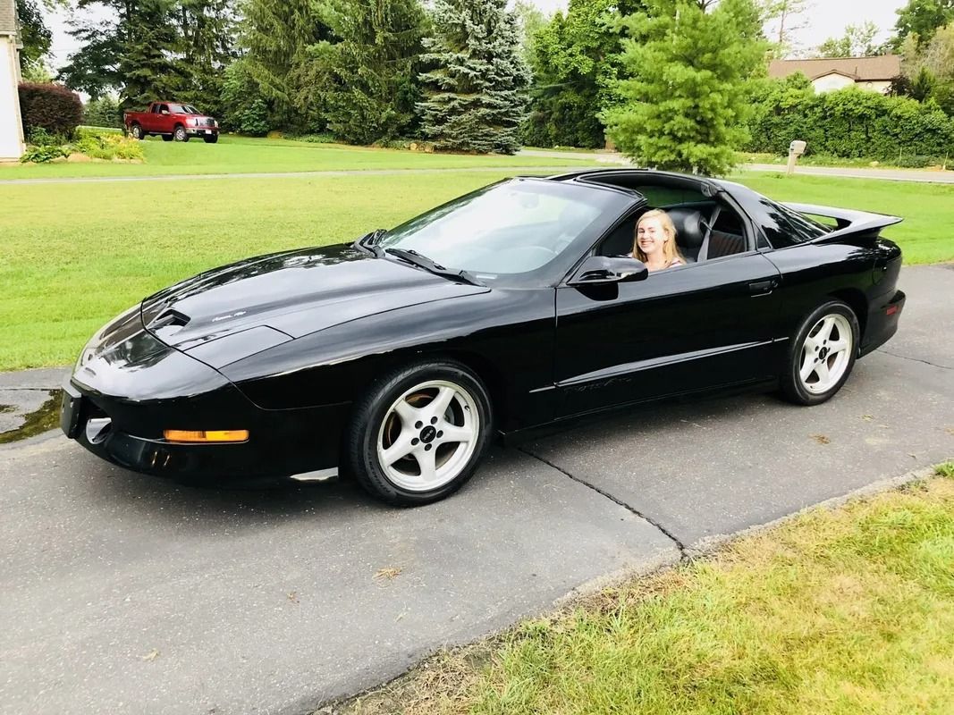 Black sports car on a driveway with a driver inside, parked beside a green lawn and trees.