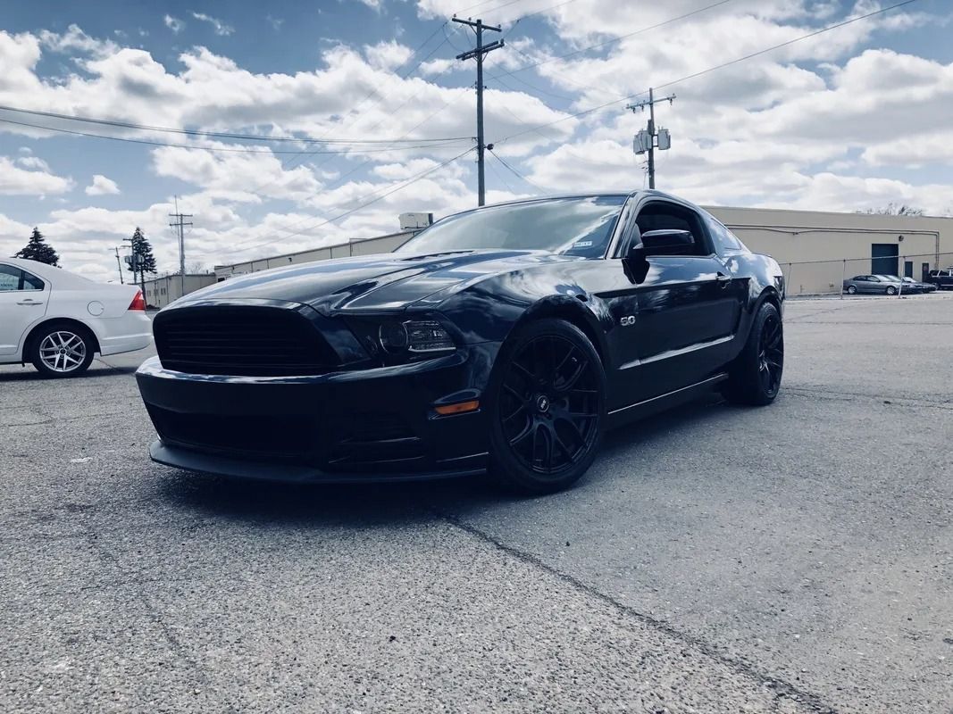 Black sports car parked on asphalt under a cloudy sky, with a white car in the background.