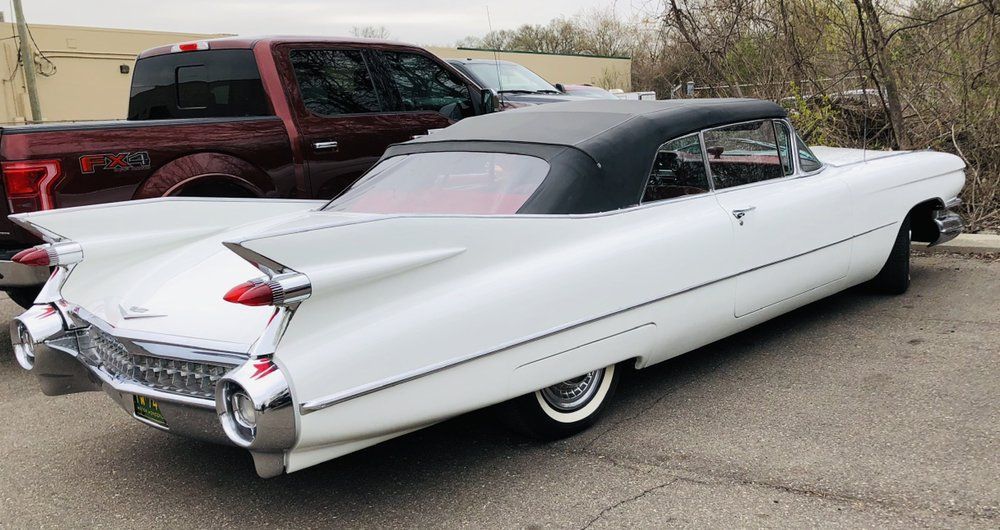 Classic white vintage Cadillac coupe parked outdoors, rear three-quarter view, with red pickup truck nearby
