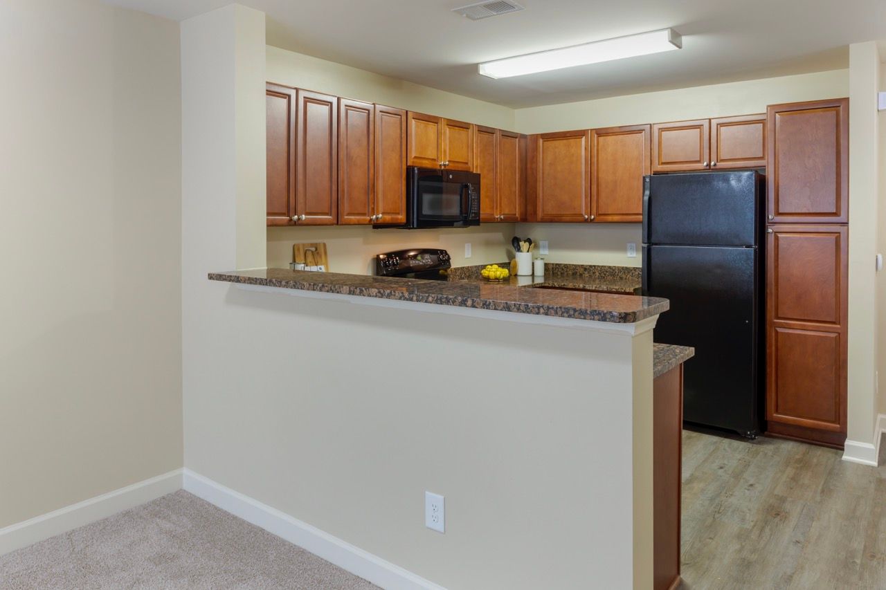 Open kitchen with brown cabinets, black appliances, and a bar counter in a beige apartment unit at Stone Gables.