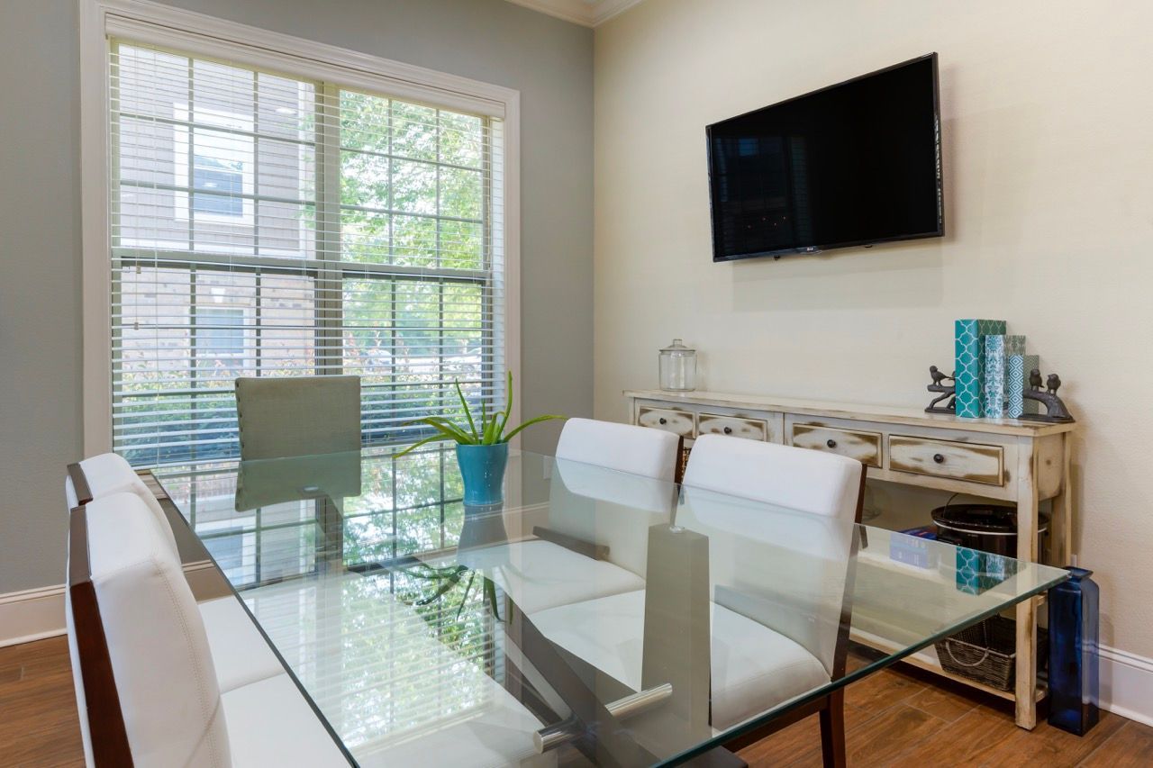 Dining area in an apartment with a glass table, white chairs, a large window, and a wall-mounted TV at Stone Gables.