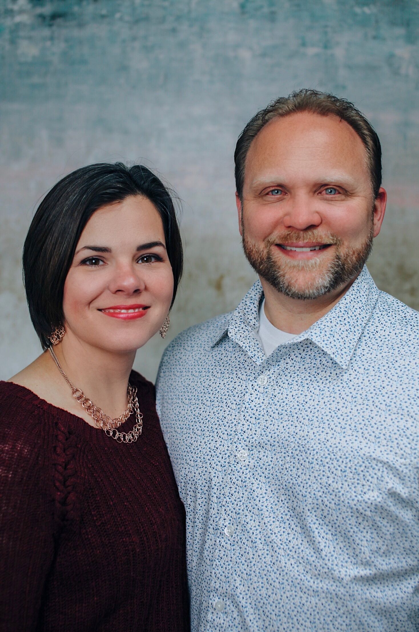 Woman with dark hair and man with beard smiling side-by-side in front of a mottled background.