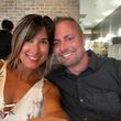 A smiling couple at a restaurant. Woman in floral top, man in black shirt. Brick wall in background.