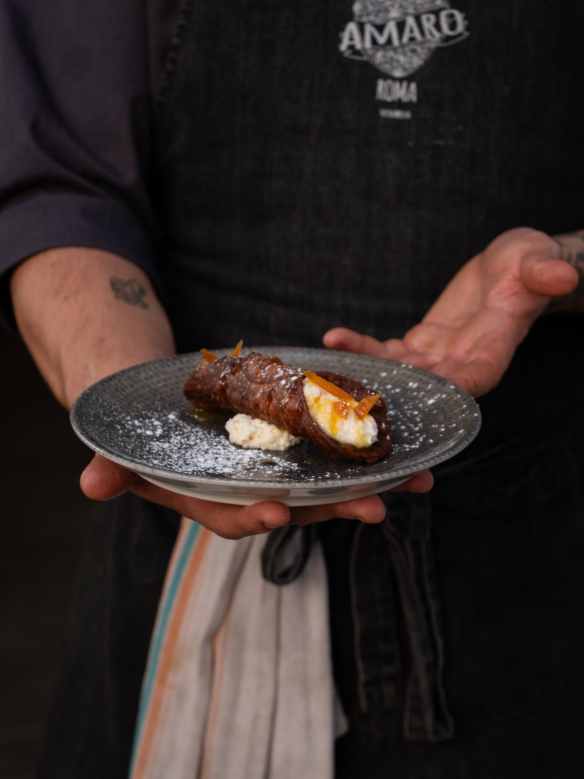 Chef che tiene in mano un piatto con un cannolo, guarnito con zucchero a velo e scorza d'arancia.