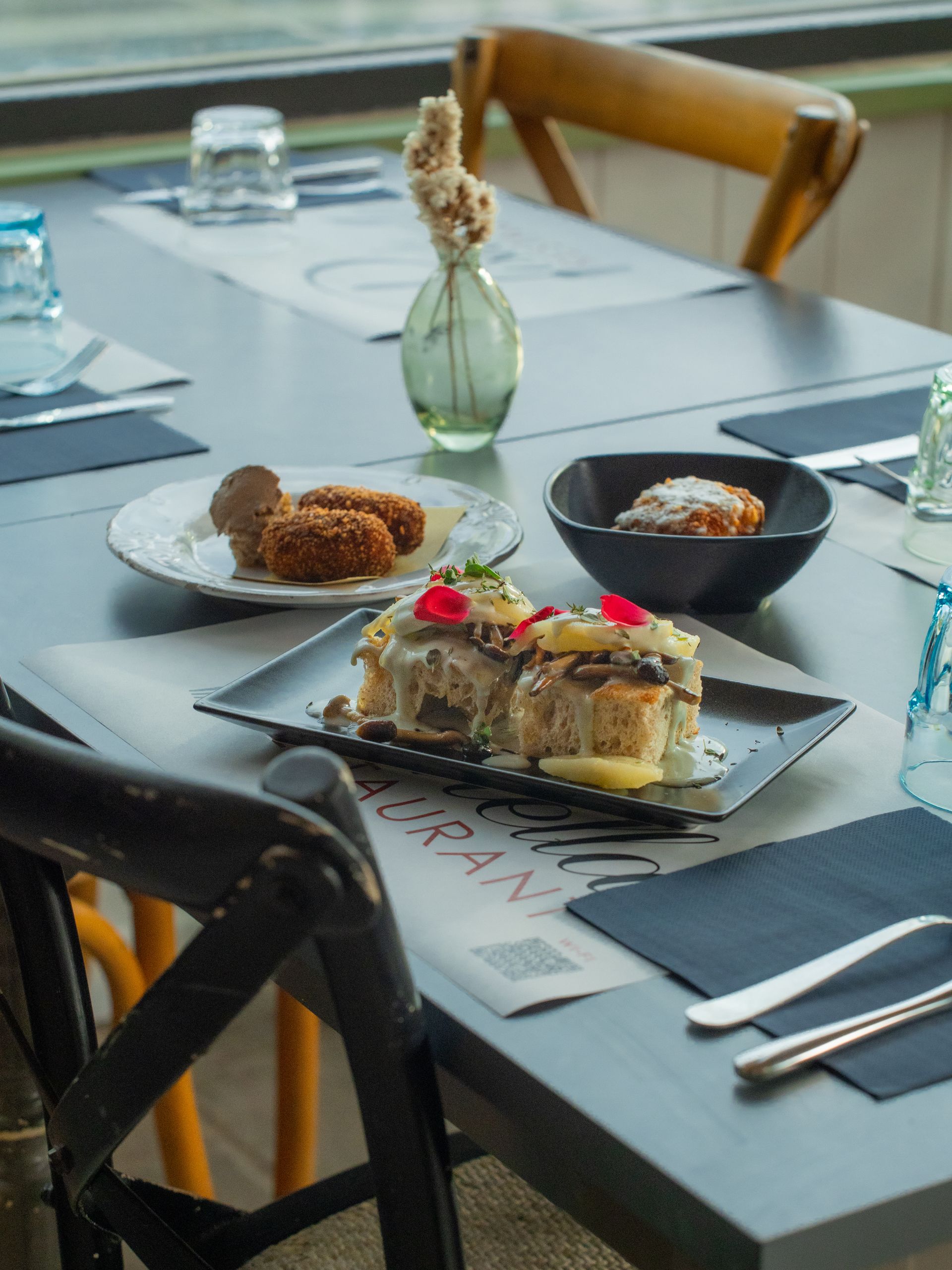 Tavola apparecchiata con piatti, tra cui crocchette e panini. Un piccolo vaso contiene fiori secchi.