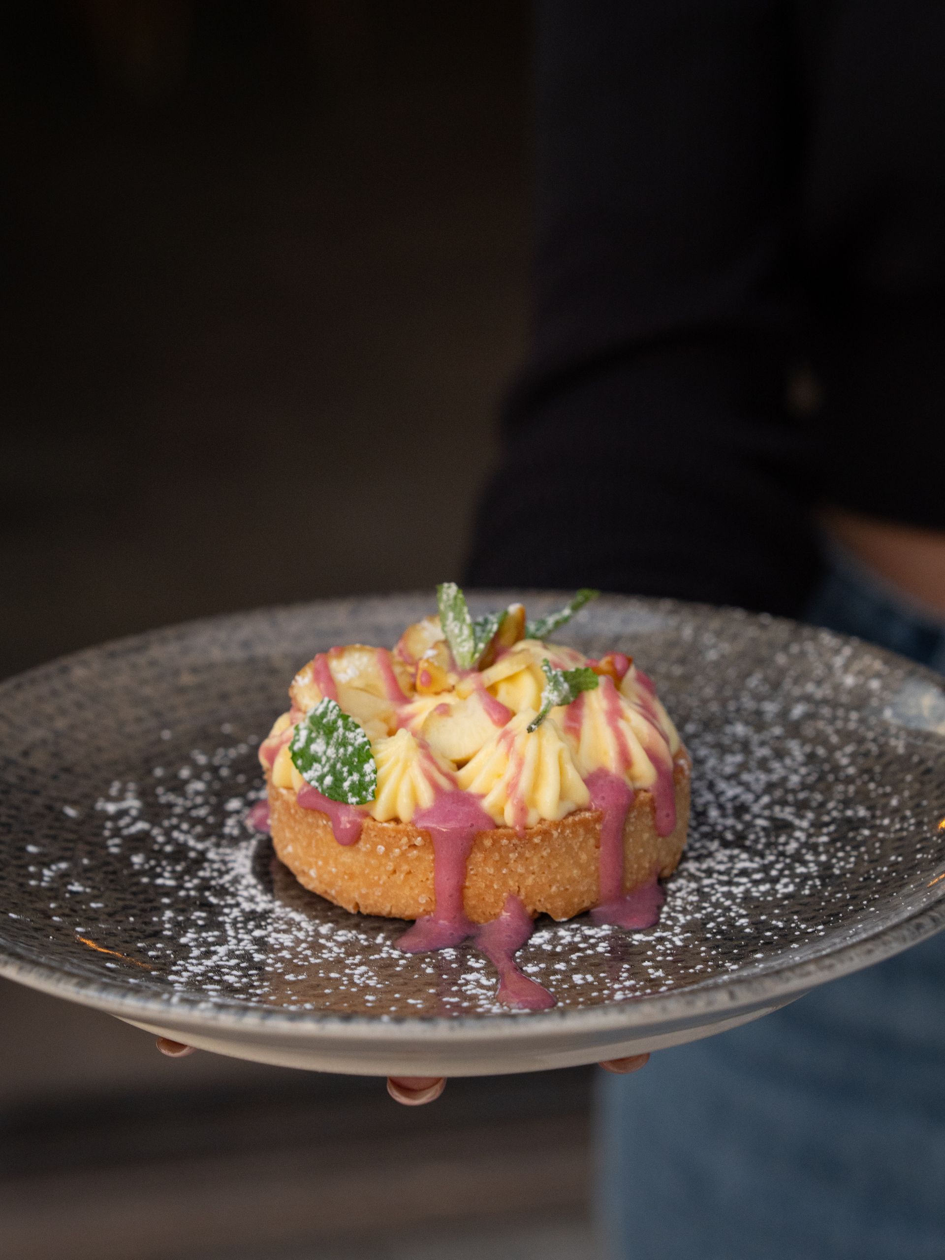 Dolce su un piatto, tenuto da una persona. Il dessert è composto da crema gialla, glassa viola e menta.