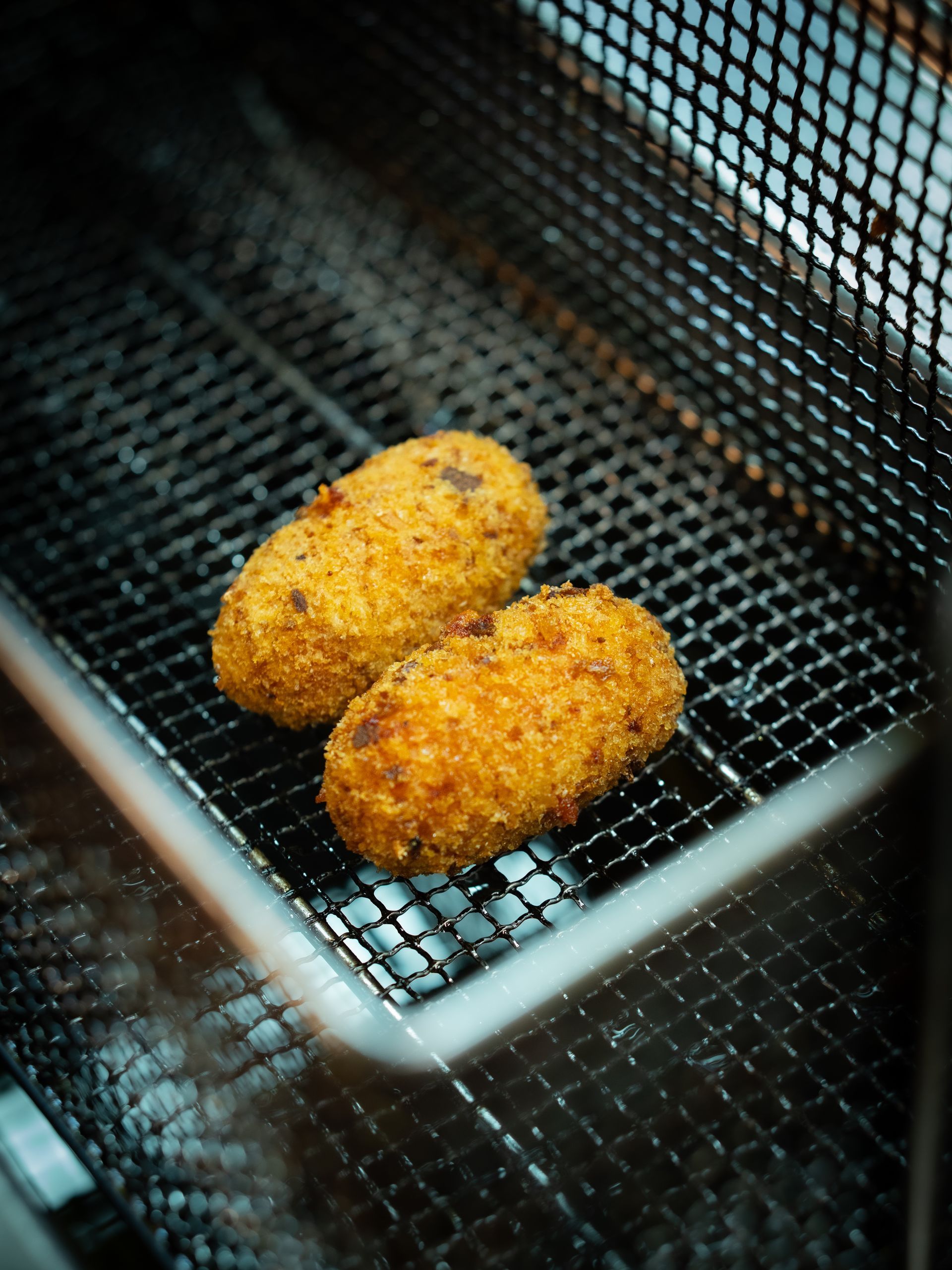 Due crocchette fritte e dorate, adagiate in un cestello di rete metallica all'interno di una friggitrice.