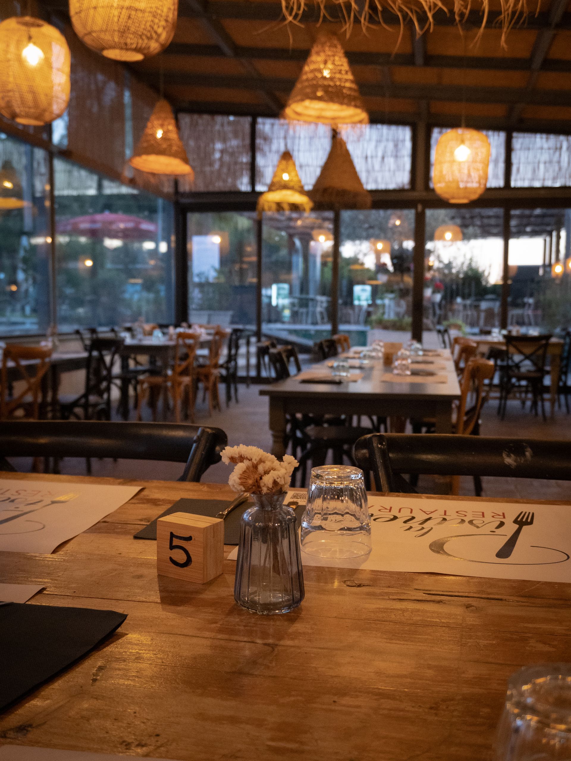 Interno del ristorante con tavoli in legno, lampade in vimini e grandi finestre che si affacciano sull'area esterna.