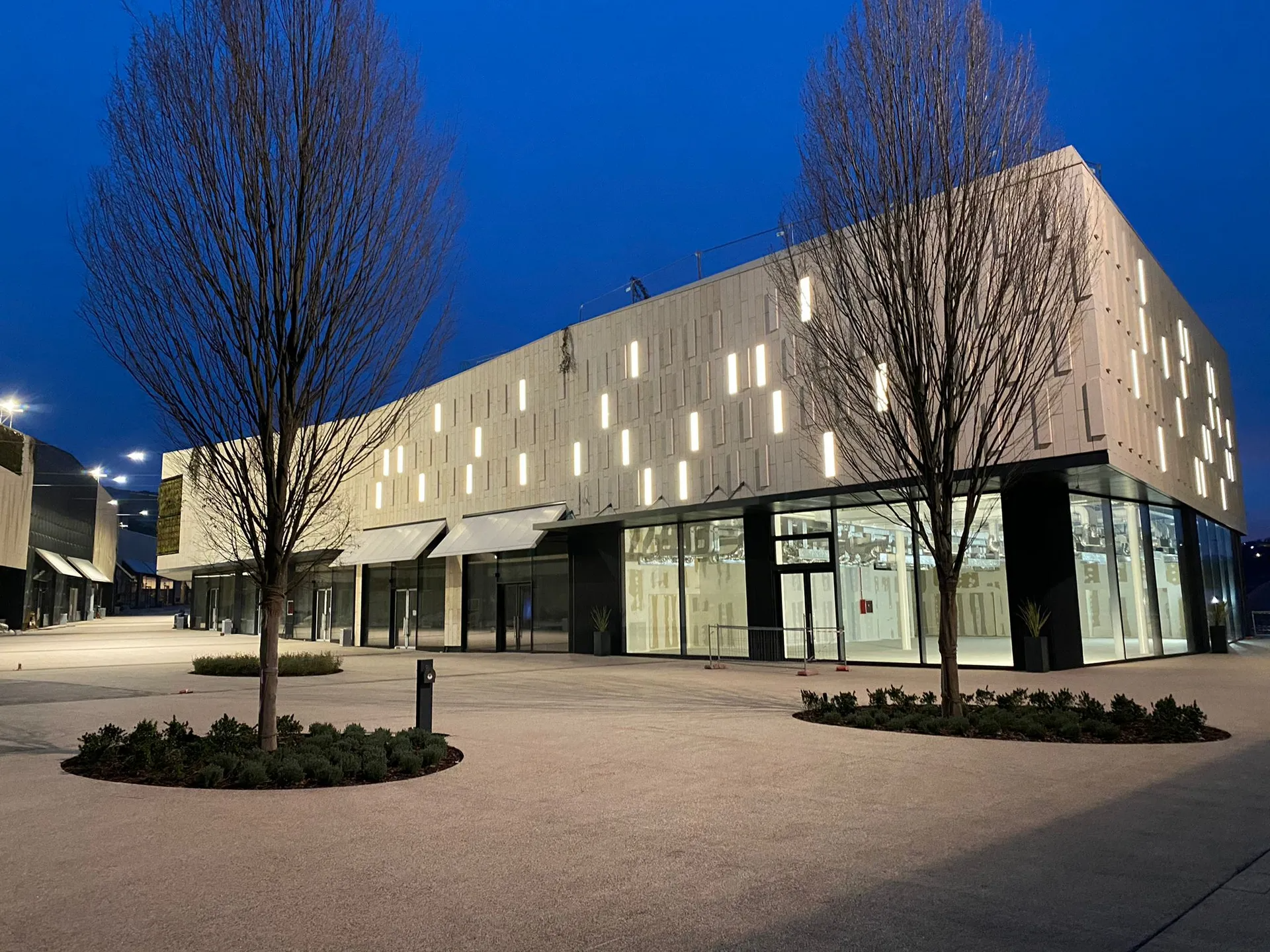 Moderno edificio commerciale con vetrine in vetro, applique rettangolari illuminate e alberi in una piazza al tramonto.