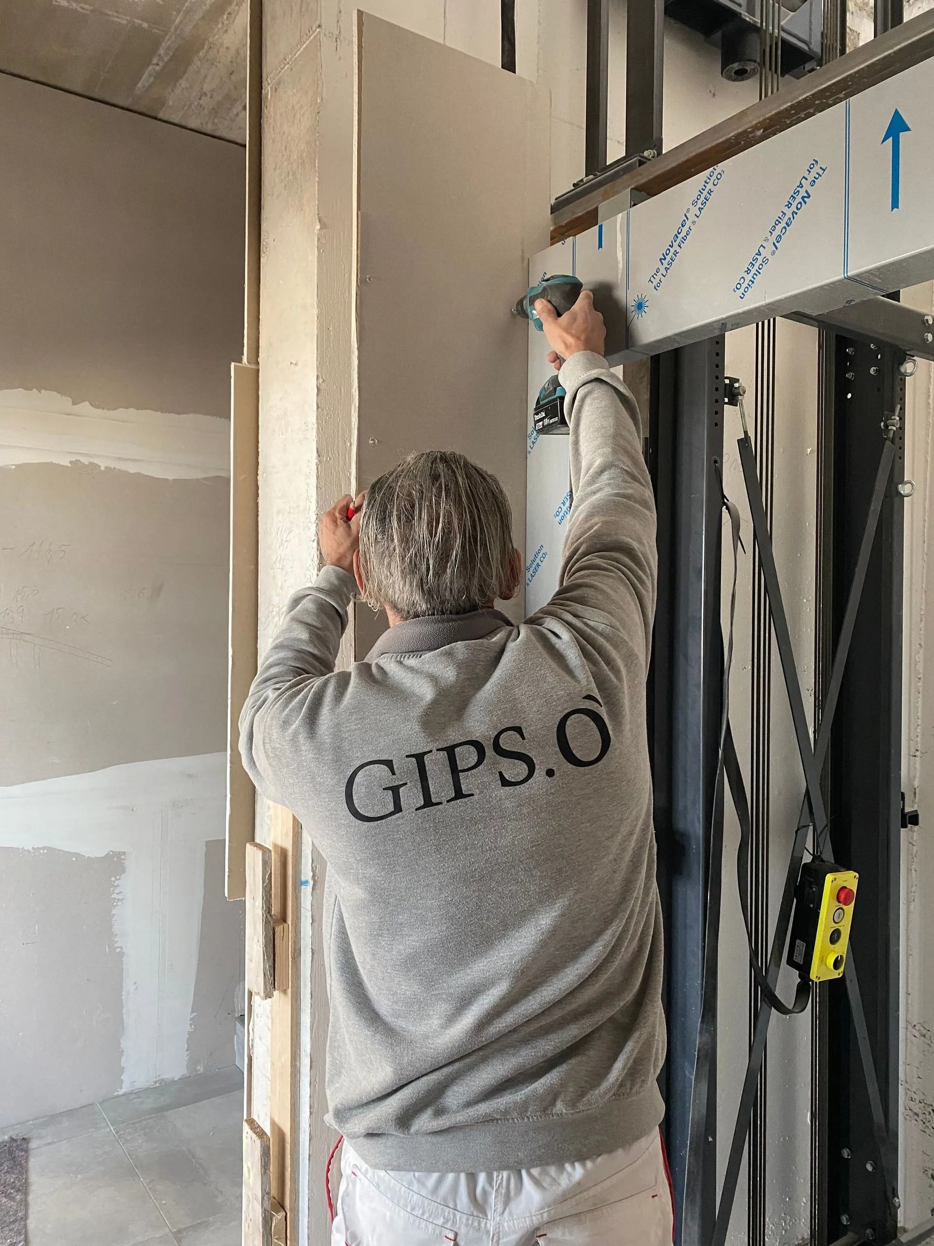 Persona che installa cartongesso utilizzando un ascensore, indossando una felpa grigia con la scritta