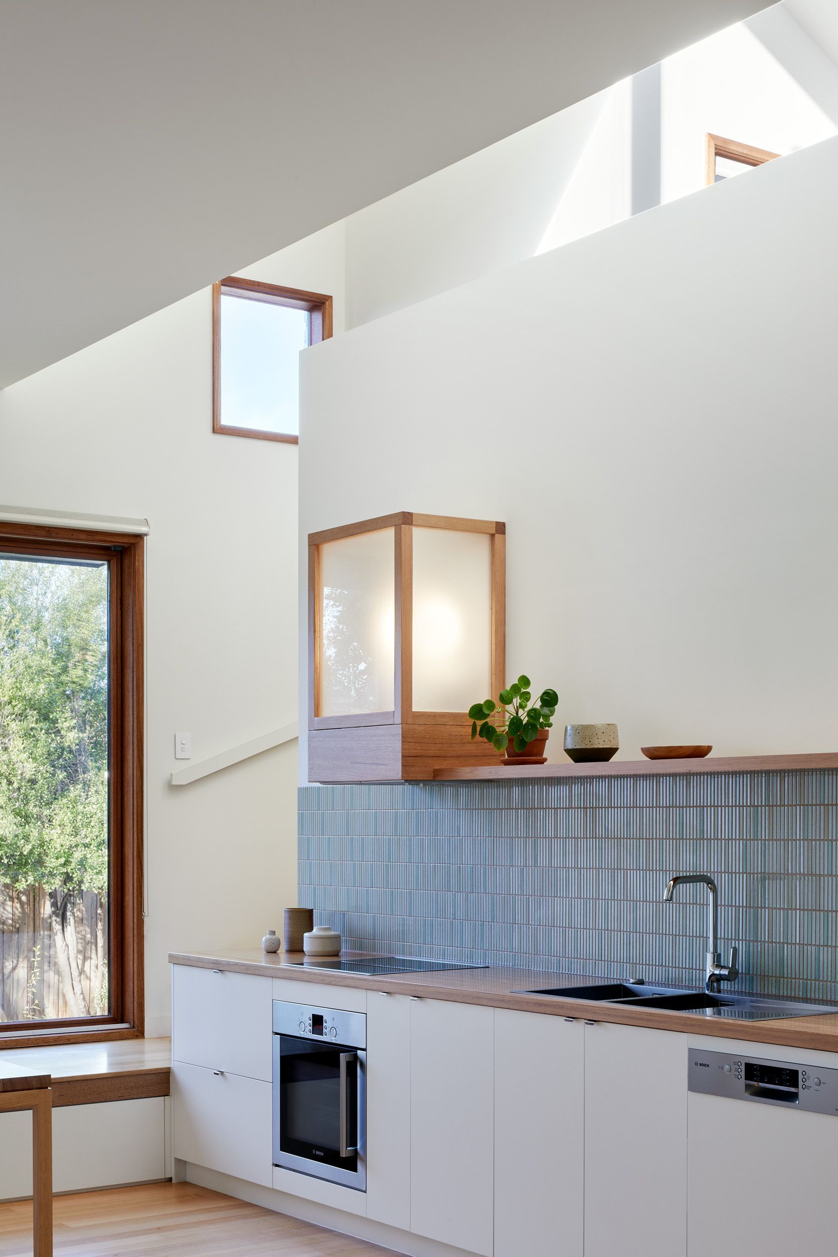 Modern kitchen with white cabinetry, kitkat tiled backsplash, and a wooden light fixture. Tall, white walls and angled ceilings with small windows.