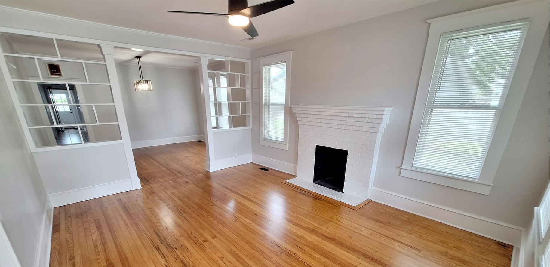 Bright empty living room with hardwood floors, white walls, fireplace, and built-in shelves, lit by large windows