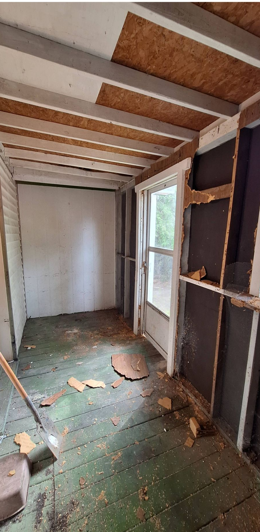 Narrow unfinished room with exposed beams, white walls, green floor, debris, and a window on the right.