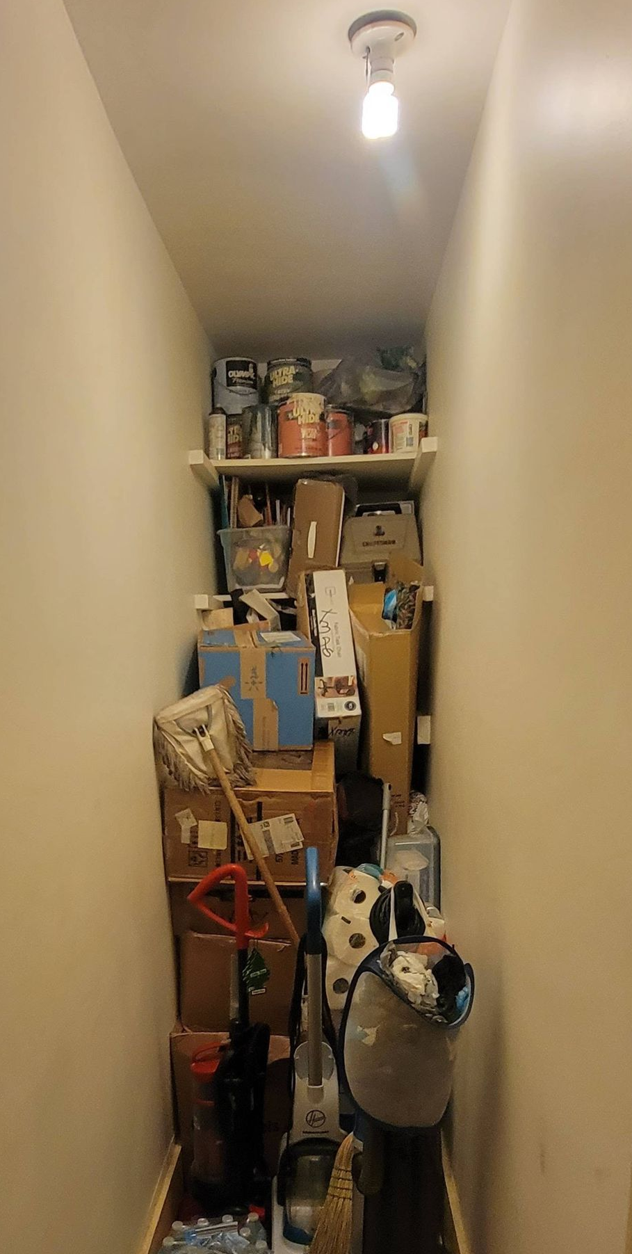 Narrow hallway cluttered with boxes, bags, and household items under a single ceiling light.