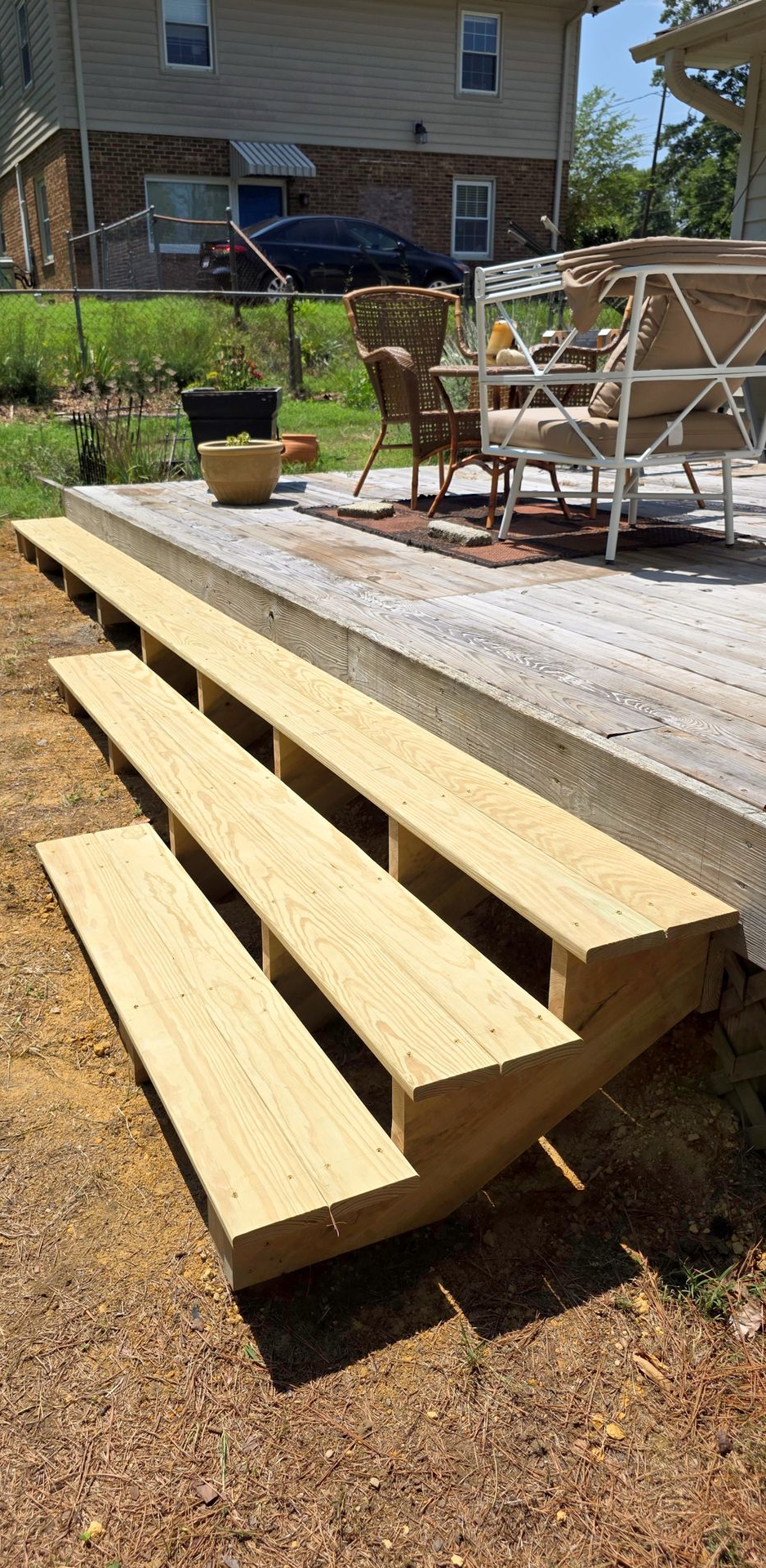 Wooden deck steps leading to a raised patio with outdoor chairs and a table in a backyard
