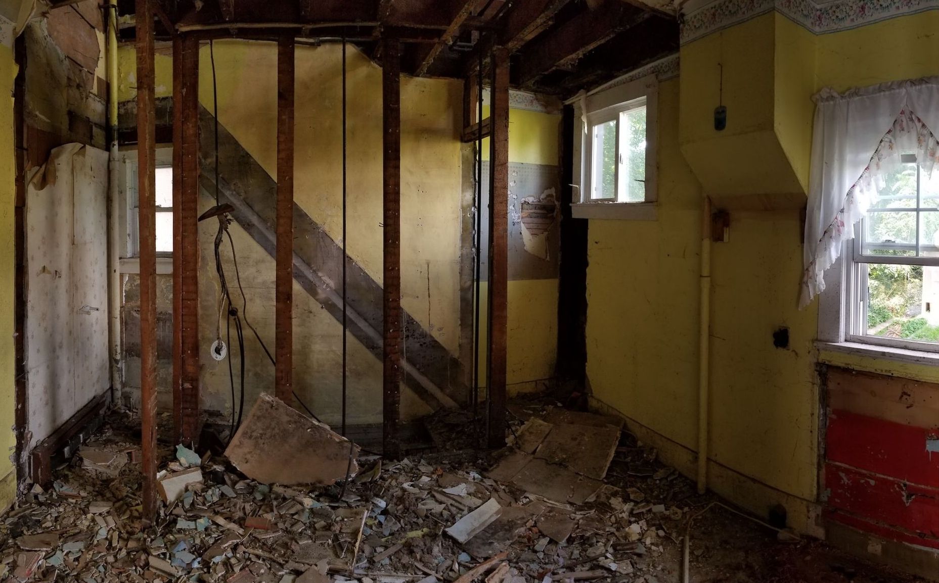 Abandoned room with damaged walls, exposed beams, debris-covered floor, and broken windows.