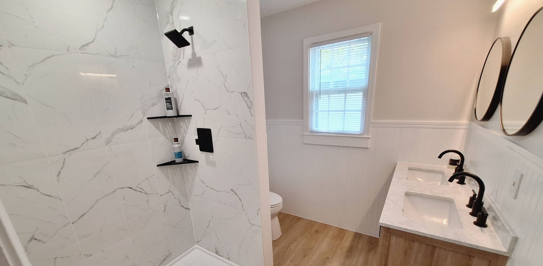 Modern bathroom with marble shower, white vanity, wood floor, and round mirrors