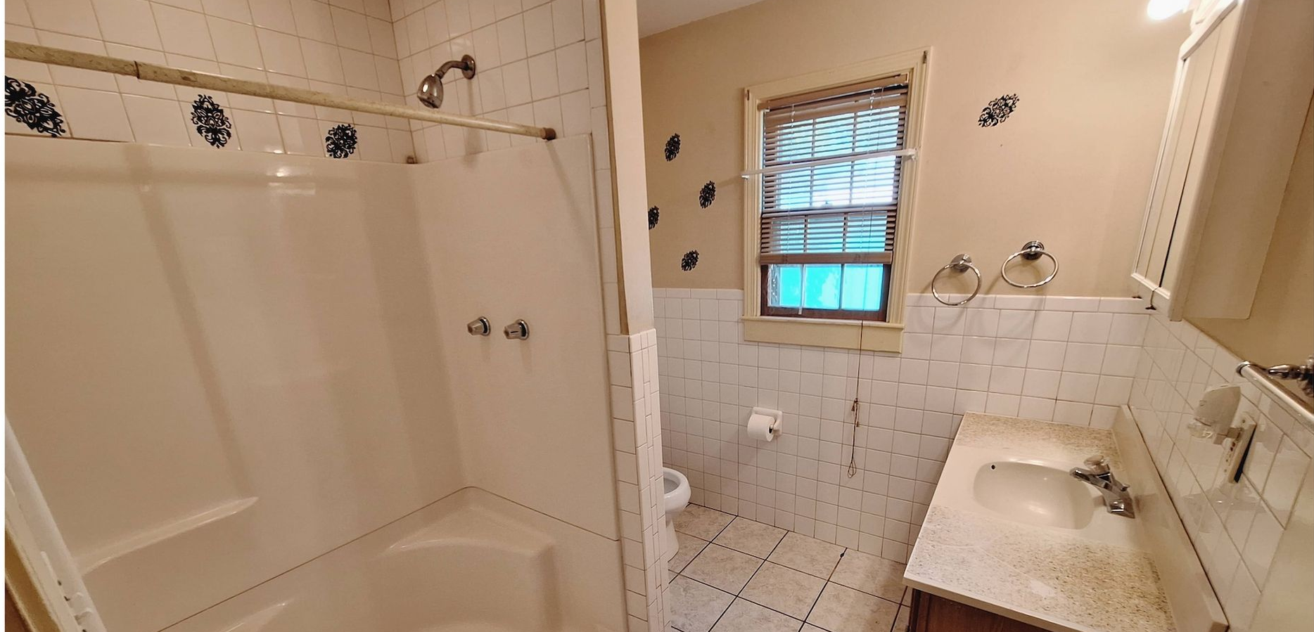 Small bathroom with a shower, toilet, sink, white tile, and a window with blue curtains.